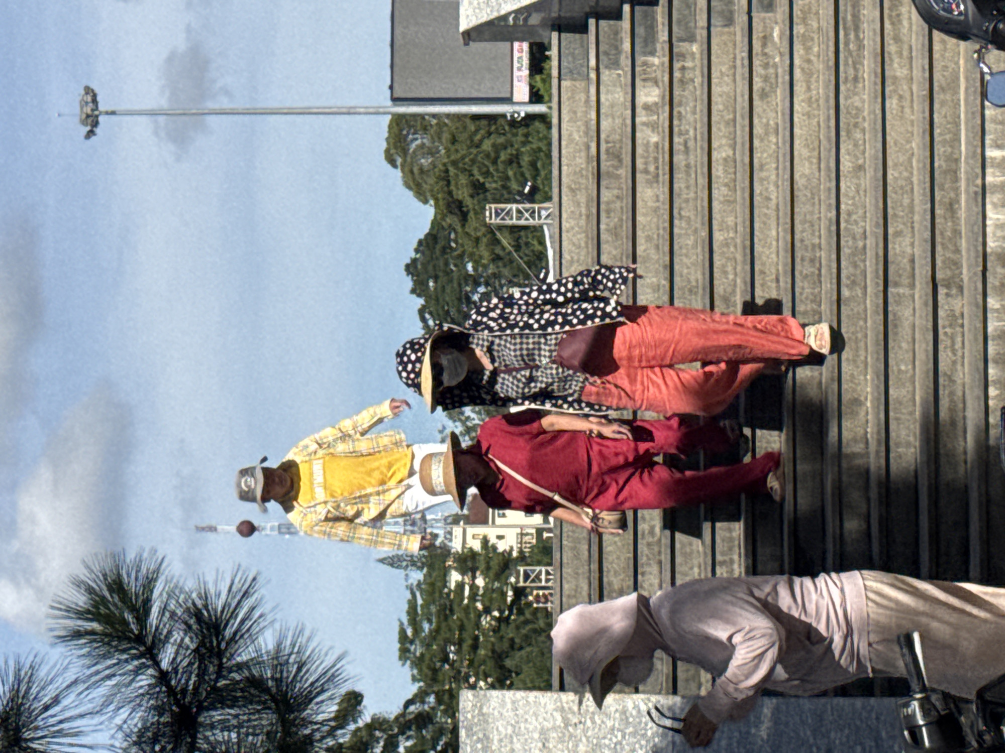 In the city of Da Lat, Vietnam, four individuals are observed walking up a wide, outdoor staircase. On the left, a person clad in a light pink hooded tracksuit and matching pants, is holding a pair of glasses in their right hand. The person is facing left, away from the stairs. Moving up the stairs, a person in a red short-sleeved shirt and red pants, wearing a straw hat and carrying a small crossbody bag, is walking up. Next to them, another person in a black and white polka dot jacket over a checkered shirt, red pants, a patterned hat, and a face mask, is also ascending the stairs, carrying a maroon shoulder bag. Higher up the stairs, a person in a yellow t-shirt and a yellow and white plaid jacket, wearing a light brown hat, is walking. The staircase is made of concrete with dark stone edges. The background features lush green trees, a tall light pole, a communication tower, and a building. The sky is partly cloudy. The overall scene suggests a casual daytime outing in an urban park or public space.