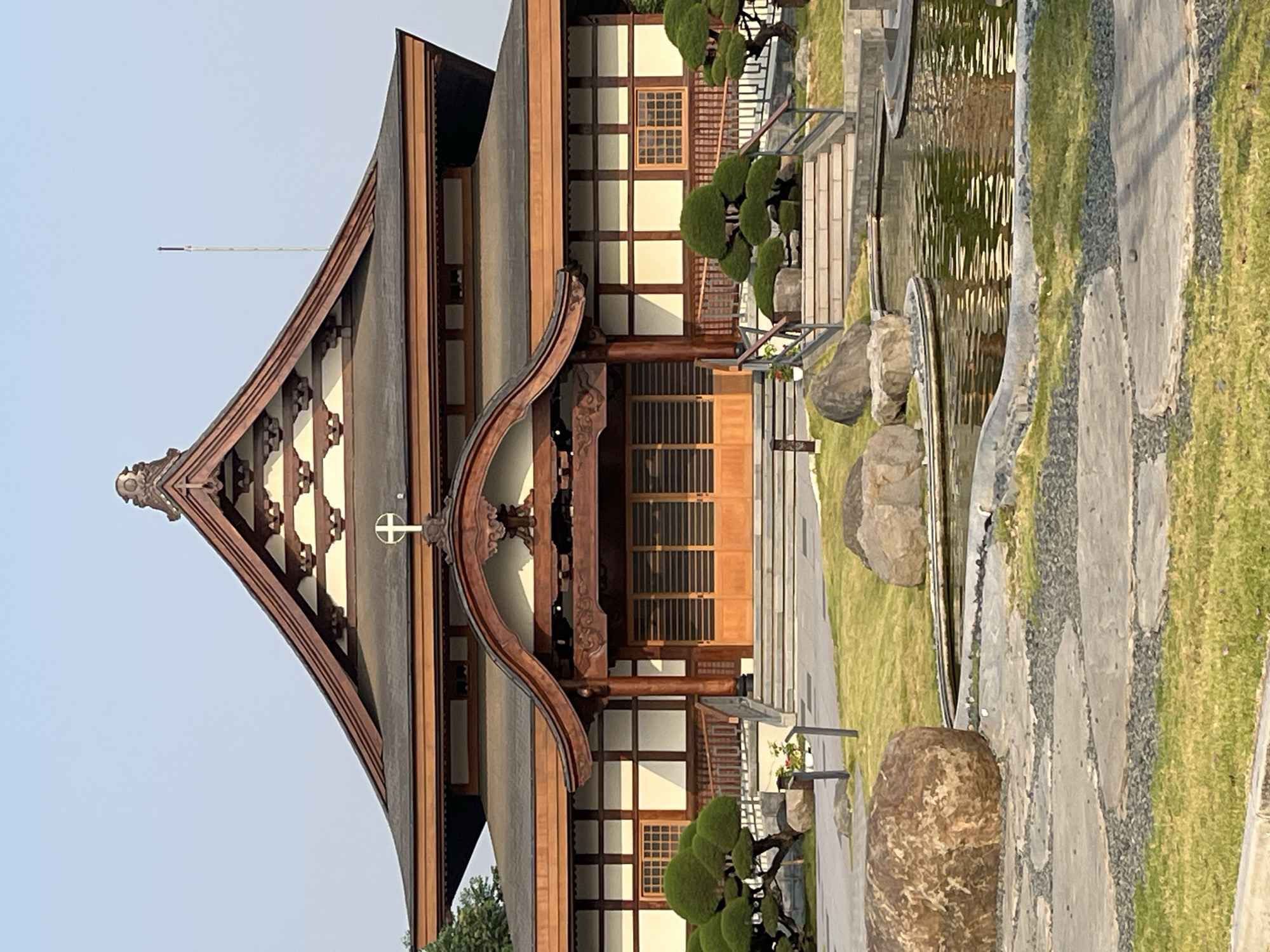 A medium shot captures the facade of a building with a Japanese architectural style, bathed in the warm, soft light of what appears to be late afternoon or early morning. The building, constructed primarily of wood and with a dark, tiled roof, features prominent eaves and intricate wooden detailing. A cross is visible at the apex of the main roof structure, suggesting a religious or spiritual affiliation.
In the foreground, a serene water feature or pond reflects the sky and surrounding greenery. The edge of the pond is lined with sculpted stones and gravel pathways, interspersed with neatly manicured grass areas. Several large, natural boulders are strategically placed within the landscape, adding to the naturalistic aesthetic.
The building's entrance is a large set of double wooden doors, flanked by wooden pillars and a wide, curved awning. To the left and right of the entrance, ascending staircases with wooden railings lead up to the building's main level. On these landings, and in the surrounding garden, are meticulously shaped bonsai trees, their dense green foliage contrasting with the warm tones of the building. The sky above is a clear, pale blue, with a tall, thin pole or antenna visible in the upper right quadrant. The overall impression is one of tranquility and cultural fusion.