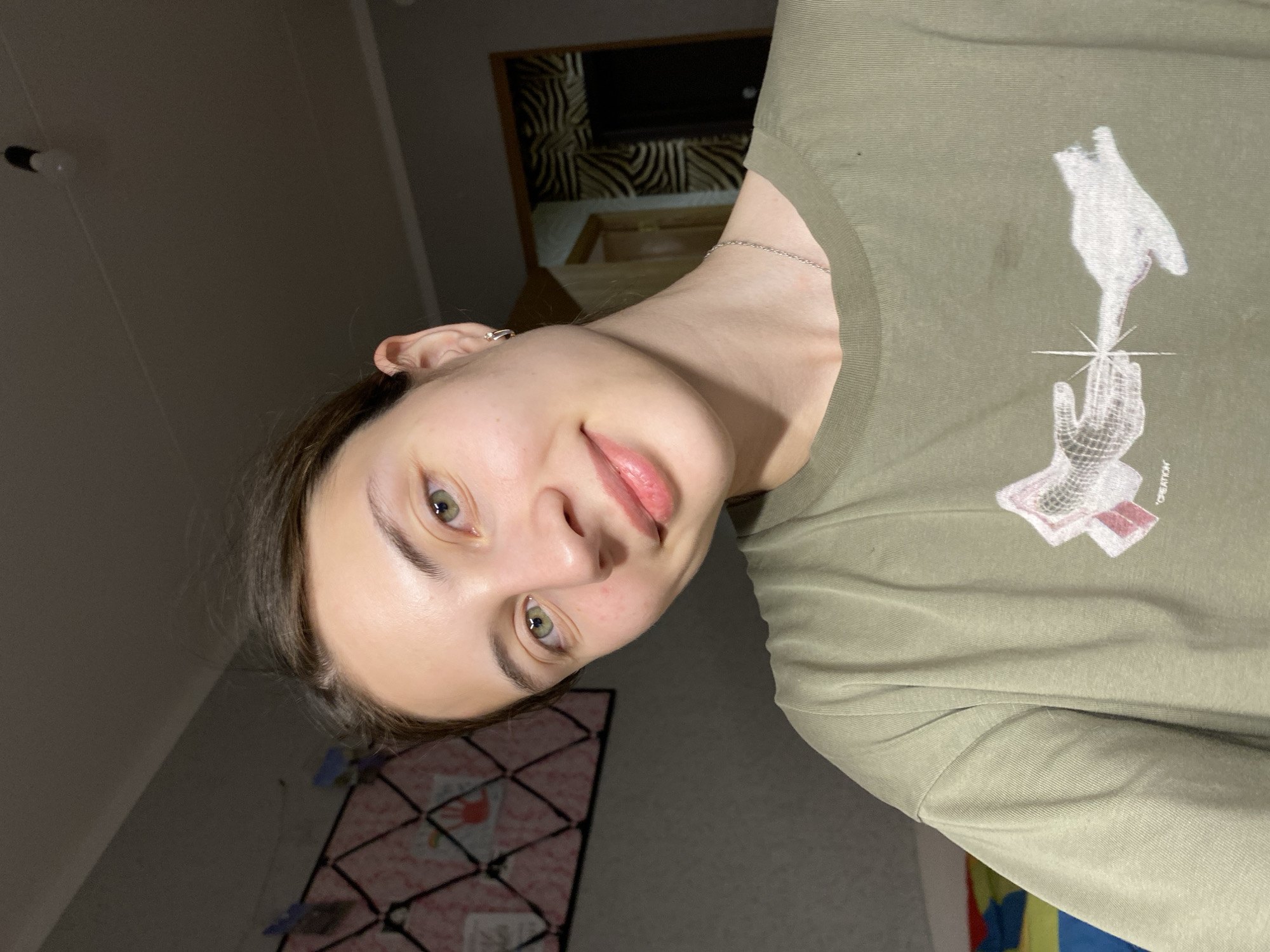 The image shows a young woman with light skin and green eyes. She has dark brown hair pulled back and is wearing a light green t-shirt with a graphic design on the front. She is smiling slightly and looking towards the camera. The background is dimly lit and appears to be an indoor setting, possibly a room. There is a bulletin board with some items pinned to it on the left side of the image, and a doorway with a patterned wallpaper on the right. A light bulb is visible on the ceiling. The overall impression is a casual, indoor portrait. The image does not provide any specific information about the location in the city of Votkinsk, Russia.