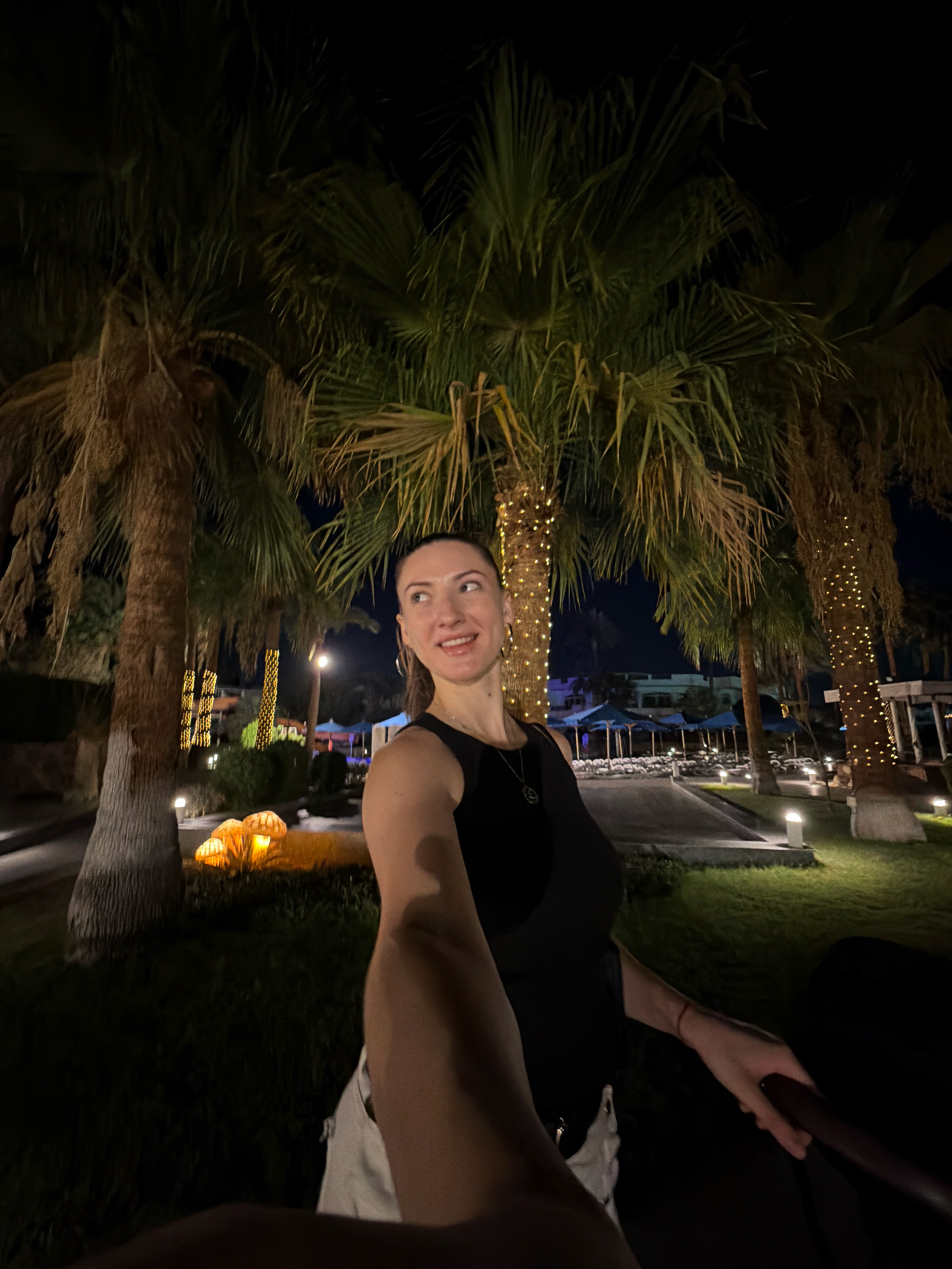 A night-time exterior shot taken in Sharm El Sheikh, Egypt, features a young woman posing for a selfie. She is wearing a black tank top and white shorts. The woman is standing in front of several palm trees, some of which are adorned with string lights. In the background, there are lounge chairs, umbrellas, and some illuminated mushroom-shaped decorations. The ground is covered in grass and pathways.