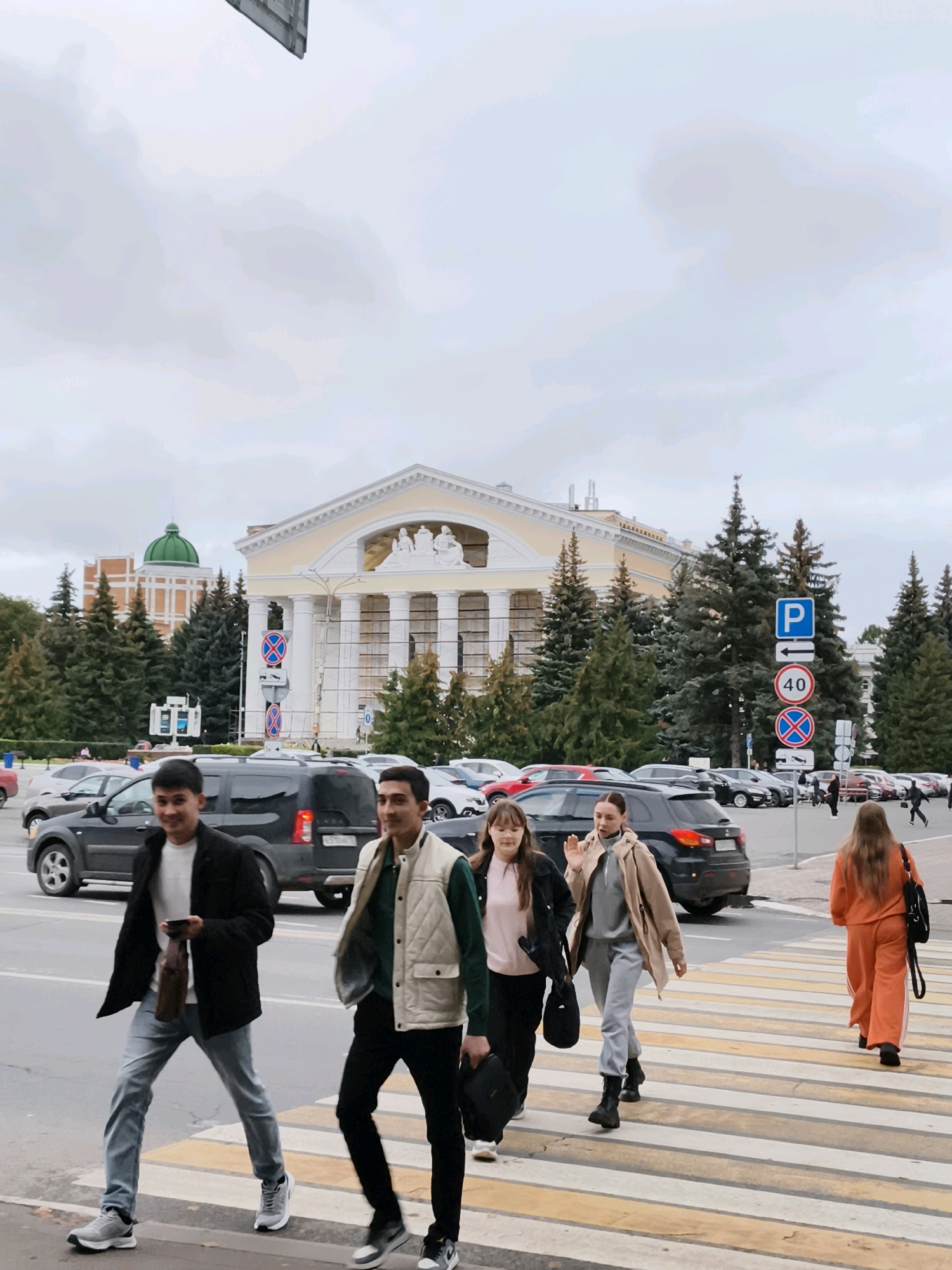 In Yoshkar-Ola, Russia, on a cloudy day, several people are crossing a pedestrian walkway in front of a large, light-yellow building with a portico supported by columns. The building appears to be a significant public structure, possibly a theater or government building, with classical architectural elements. Behind the building and the people, a grove of mature evergreen trees stands tall. Several cars are visible, including black SUVs and other passenger vehicles, suggesting a busy urban environment. Traffic signs, including parking and speed limit signs (40), are posted near the roadside. The people are dressed in casual, modern clothing. In the foreground, four individuals are walking across the zebra crossing. On the left, two young men are walking; one is wearing a black jacket and jeans, holding a dark brown bag. The other is wearing a light-colored vest over a green shirt and black pants. To their right, two young women and another woman are crossing. One woman in a pink shirt and black jacket is walking with a woman in a beige jacket and grey tracksuit, who appears to be gesturing. Further to the right, a woman in an orange tracksuit is walking away from the camera. The overall atmosphere is that of a typical day in a city square.