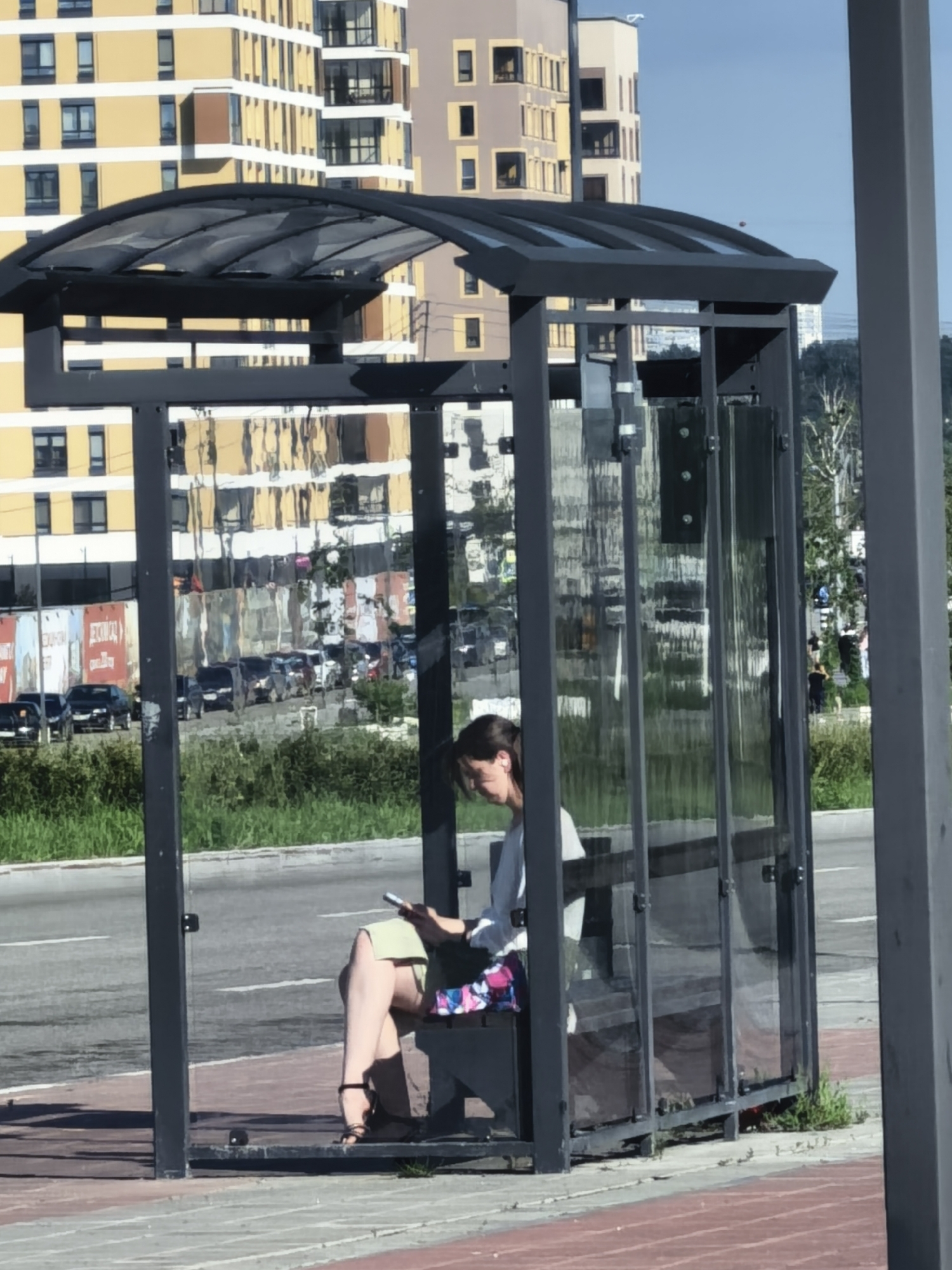 A woman sits inside a glass-enclosed bus shelter in a city. She is wearing a light-colored shirt and a light green skirt, and is looking at her phone. Her legs are crossed, and she is wearing black sandals. The bus shelter has a dark metal frame and a curved, transparent roof. Behind the shelter, a street with cars is visible, and beyond that, multi-story buildings with yellow and brown facades. Green grass and trees are present in the foreground and background. The sky is blue with some wispy clouds.