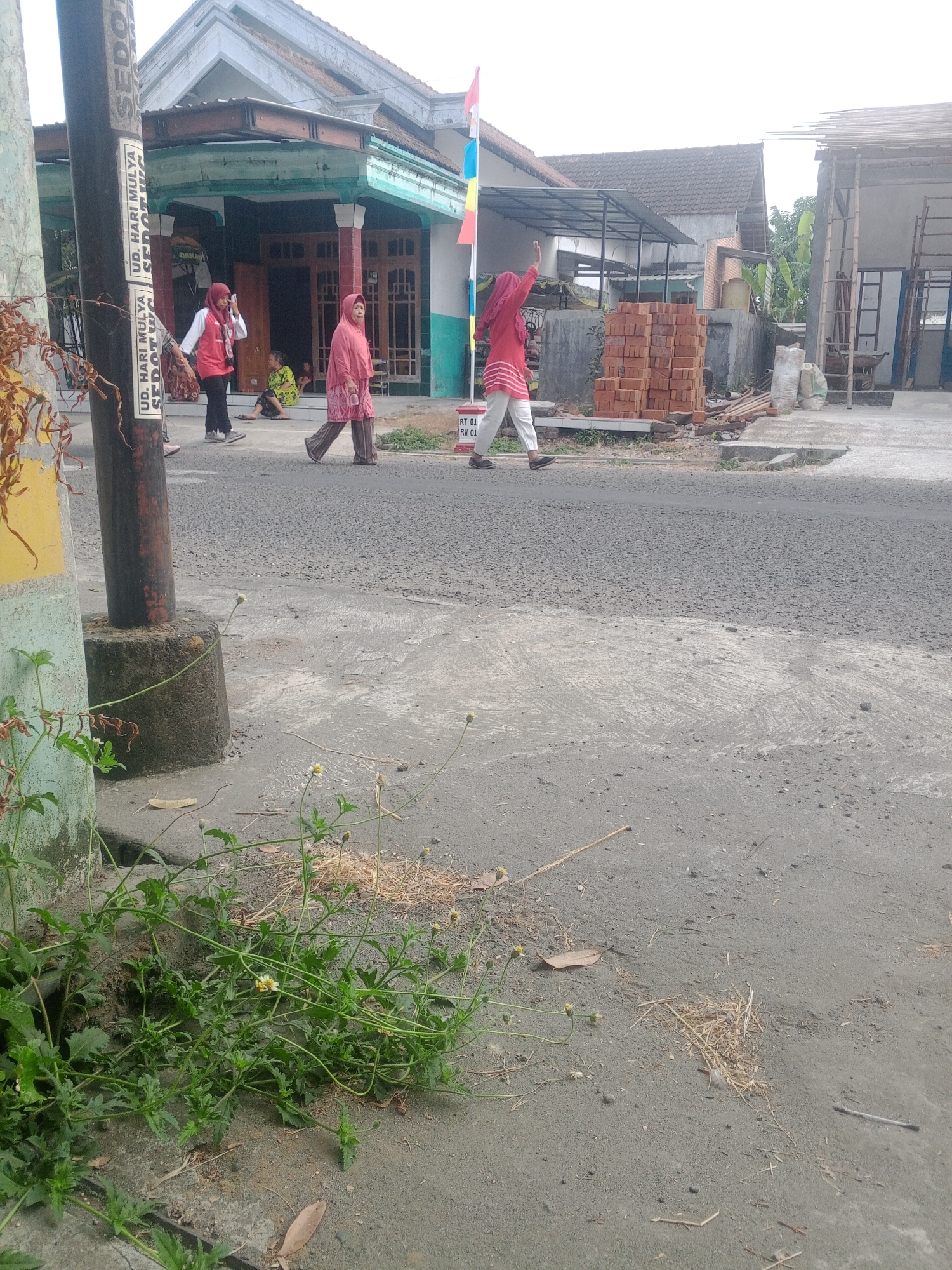 The image depicts a street scene in Lembah, Indonesia. In the foreground, a concrete and asphalt road is visible, with a concrete post on the left side of the frame. Sparse green vegetation with small white flowers grows along the edge of the road. In the middle ground, a paved street runs horizontally. Three individuals are walking on the street. On the left, a person in a red top and black pants, wearing a headscarf, walks away from the camera. To their right, a person in a pink headscarf and patterned pink and brown clothing walks towards the camera. Further to the right, a person in a red top and white pants, also wearing a headscarf, is walking away from the camera and waving their right hand. Behind them, a stack of red bricks and construction materials are visible, along with a partially constructed building. In the background, several houses with tiled roofs are present. A flag pole with Indonesian flags is also visible near the houses. The environment appears to be a residential area with ongoing construction.