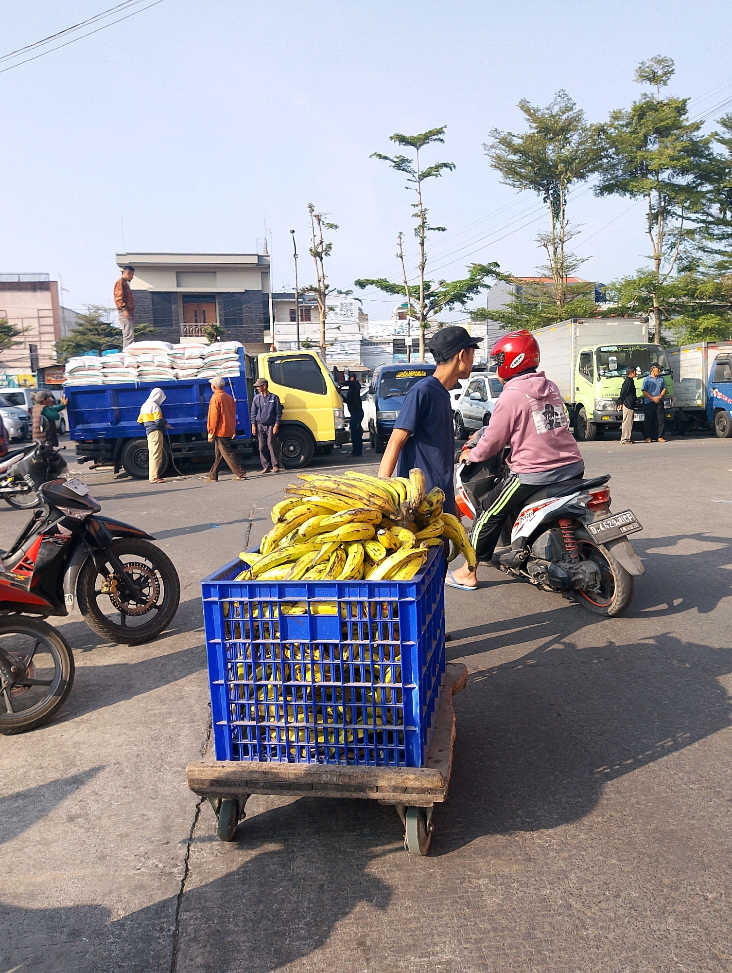 This image depicts an outdoor scene in Lembang, Indonesia, likely at a market or distribution point. In the foreground, a blue plastic crate overflowing with ripe bananas rests on a wooden cart with wheels. To the left, the front portion of a red and black motorcycle is visible, parked. In the midground, a yellow truck with a blue cargo bed, loaded with sacks of goods, is parked. Several individuals are interacting around the truck. One man in an orange shirt walks away from the truck, while another in a blue jacket stands beside it. A person in a white hooded jacket is also near the truck. Further back, a person in a pink hoodie and red helmet is seated on a motorcycle, facing away from the camera and towards the truck. Two other individuals are visible in the background near other vehicles, including a light green truck and a dark blue van. The environment appears to be a paved area with a clear sky overhead. Trees with sparse foliage are scattered throughout the scene, and buildings are visible in the background. The overall impression is one of a busy, commercial exchange taking place outdoors.