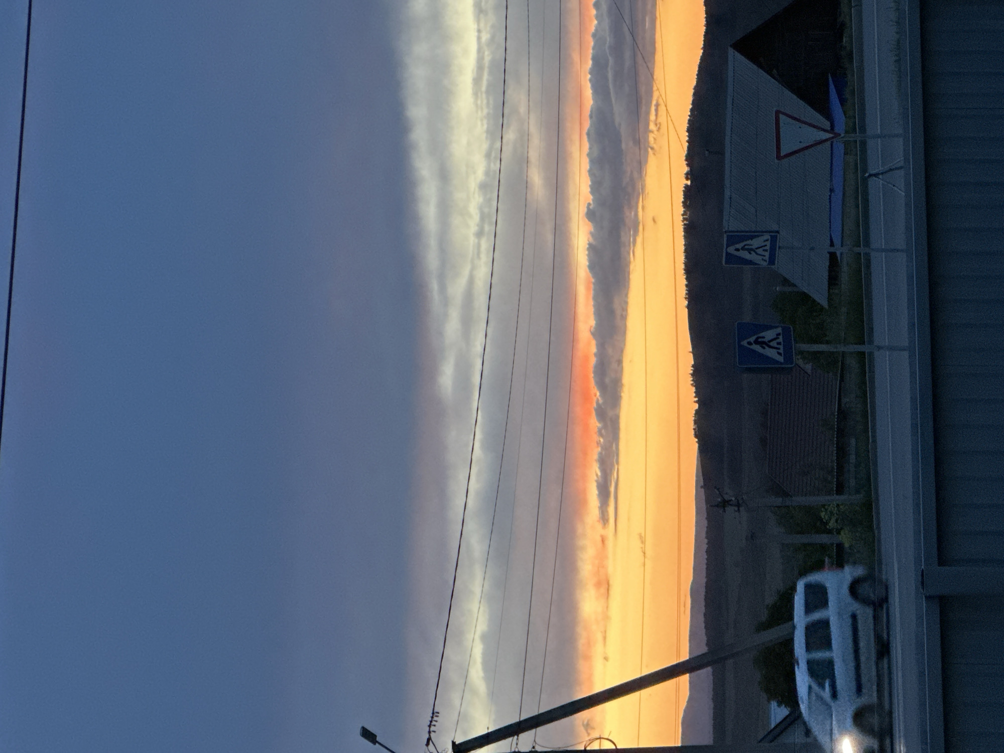 The image shows a street scene in Uchaly, Russia during a sunset. The sky is divided into two sections: a dark blue area on the left and a bright orange and yellow area on the right where the sun is setting. A vertical line of clouds separates the two sections. Power lines run horizontally across the sky. On the right side of the image, there is a street with pedestrian crossing signs and a warning sign, as well as a white van parked along the side of the road. Buildings and vegetation are visible in the background. A street lamp is also visible on the left side of the road.