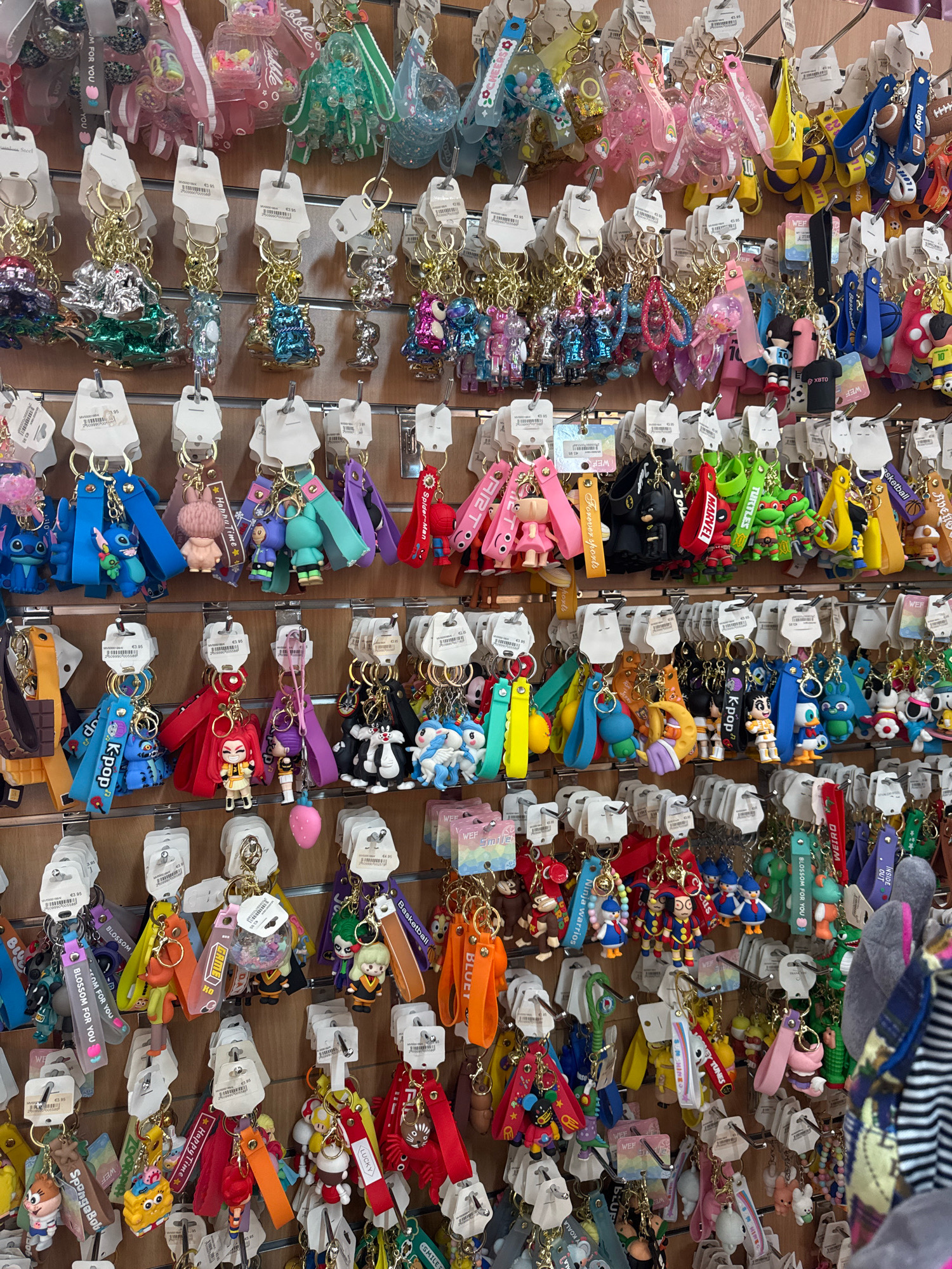 The image shows a wall display of numerous keychains in a retail setting. The keychains are colorful and feature a variety of characters, objects, and designs. They are hung on hooks attached to a wooden panel. The display is densely packed with keychains, organized in horizontal rows. The lighting appears to be artificial, casting a slight shadow on some of the items. The background is out of focus but suggests a shop environment. The location is identified as Limassol, Cyprus, implying this is likely a souvenir shop or a store selling accessories. There are no people visible in the image.