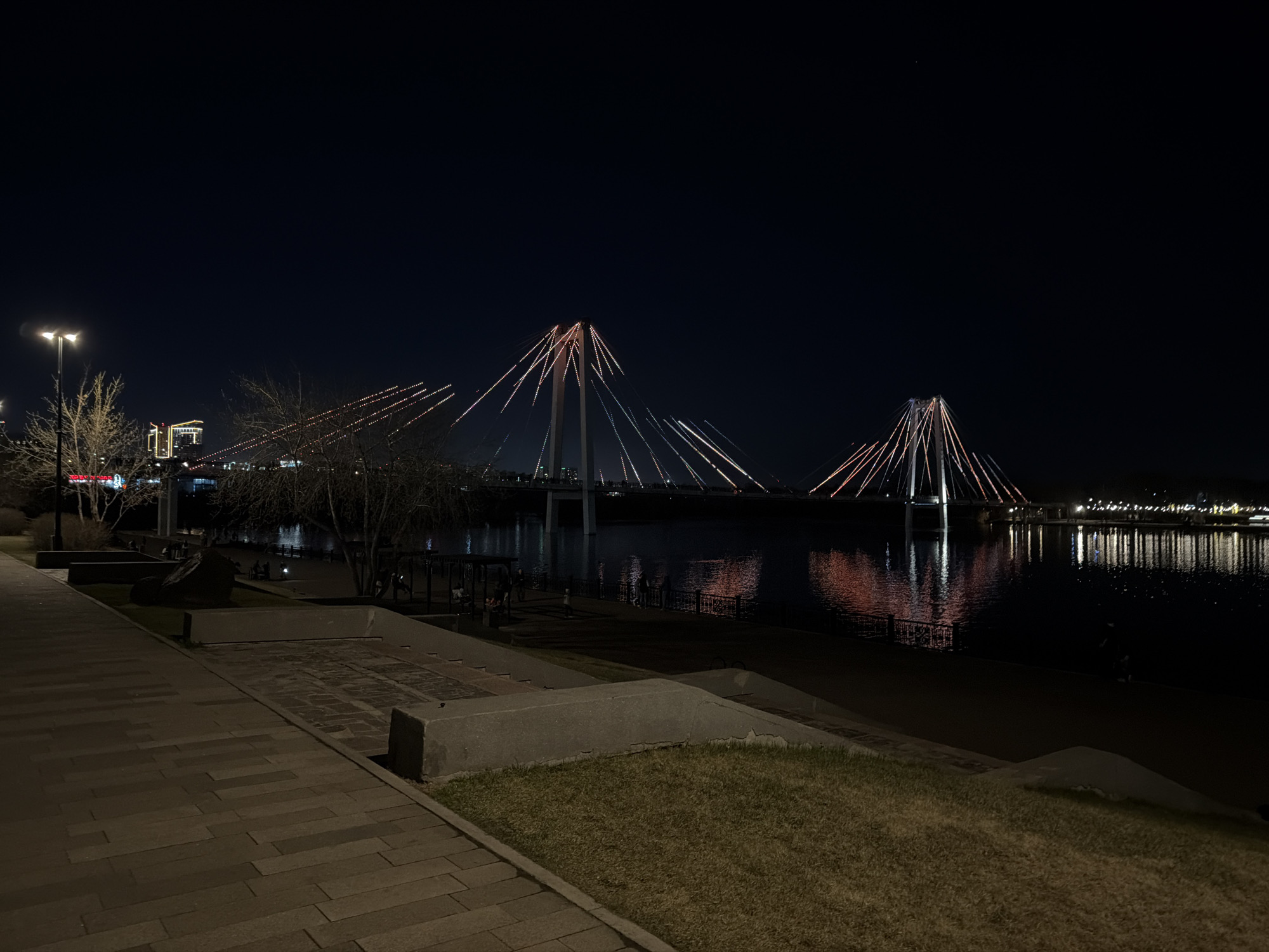 The image shows a night view of the Yenisei River in Krasnoyarsk, Russia. Two cable-stayed bridges are illuminated in the background, with their reflections visible in the water. In the foreground, there is a paved walkway with grass patches. Bare trees are seen along the riverbank. The night sky is dark, and city lights are visible in the distance.