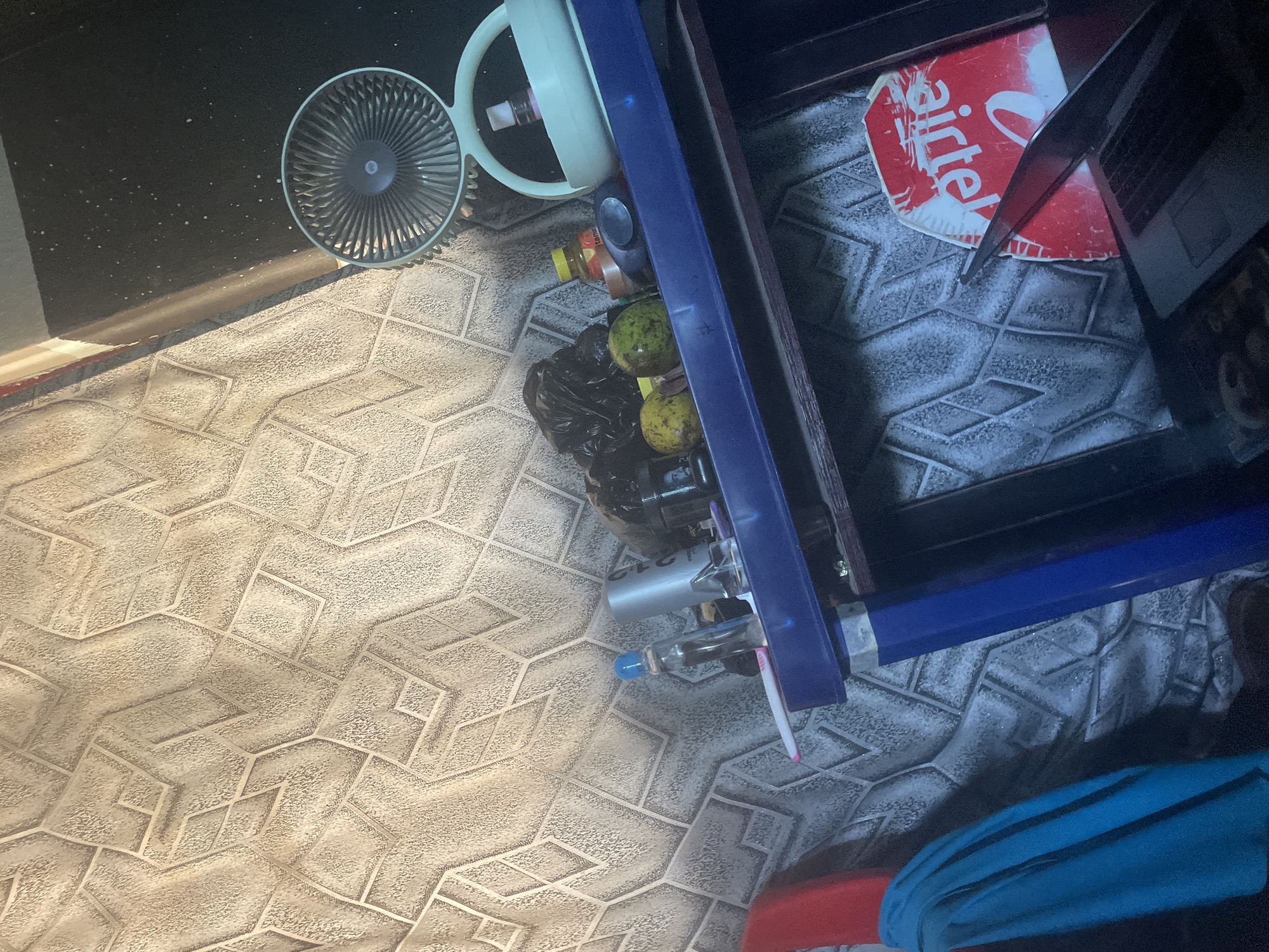 A cluttered shelf is pictured against a textured wallpaper. On the shelf, from left to right, are a bottle with a blue cap, a silver cylinder with writing, a black plastic bag, two light green fruits, a small yellow bottle with red lettering, a dark gray object, and a white fan. To the right of the fan is a white object with a handle. Below the shelf is a laptop with a red and white sticker that says "Airtel". A blue cloth hangs on the left side of the image, and a red object is partially visible behind it. The scene appears to be indoors, in what could be a home or a small shop. The image is shot from a slightly elevated angle, looking down at the shelf. The overall impression is one of disarray and everyday life.