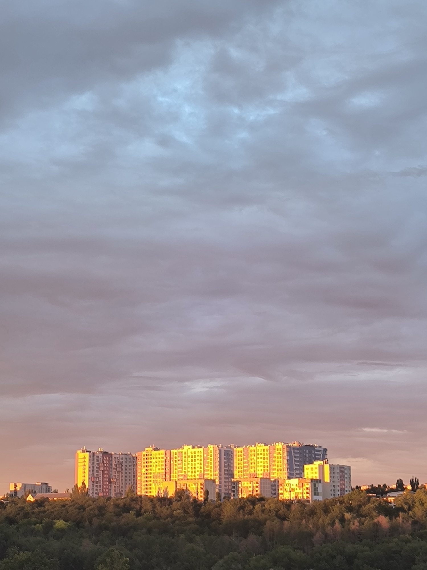 The image depicts a panoramic view of a city skyline at sunset. A dense canopy of trees occupies the foreground, with various apartment buildings rising above them in the middle ground. The buildings, particularly a large, brightly lit yellow one on the right, are bathed in the warm glow of the setting sun. The sky above is a dramatic mix of grey, pink, and blue clouds, with the sun's rays piercing through, creating a vibrant spectacle. The architecture suggests a Soviet-era urban planning style, common in Chișinău, Moldova. The overall scene is tranquil and picturesque, capturing the beauty of a city transitioning from day to night.