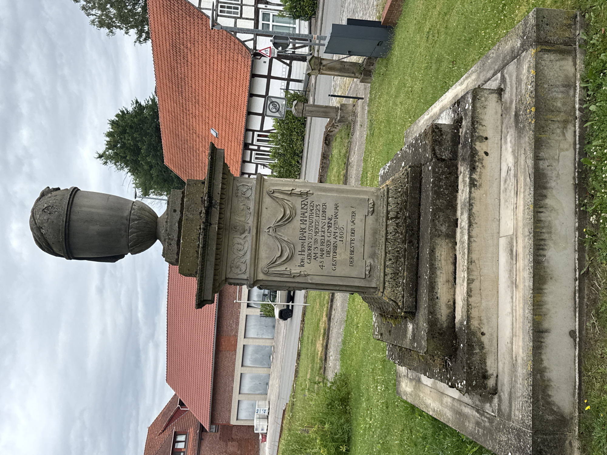 This is a photograph taken outdoors in what appears to be a historical area, possibly a cemetery or a memorial site, in Bassum, Germany. The central subject is a stone monument with an urn on top and an inscription on its base. The monument itself is weathered, with moss and discoloration visible on the stone.
The inscription on the monument is in German and reads:
"JOH: HERM: BARCKHAUSEN
GEBOREN ZU STADTHAGEN
AM 3 TEN MERTZ 1723
48 JAHR RELIGIONS LEHRER
DIESER GEMEINE,
GESTORBEN AM O TEN JANUAR
1806.
DER BESTE DER VÄTER!"
This translates to:
"John Hermann Barckhausen
Born in Stadthagen
On March 3, 1723
48 years a religious teacher
of this community,
Died on January 10,
1806.
The best of fathers!"
In the background, there are buildings with traditional German half-timbered architecture, featuring red tile roofs. The sky is overcast, suggesting cloudy or rainy weather. Green grass covers the ground around the monument. A road and a few signs, including a "30 ZONE" speed limit sign, are visible in the mid-ground, indicating the presence of a street or pathway nearby. The overall impression is one of historical significance and remembrance.