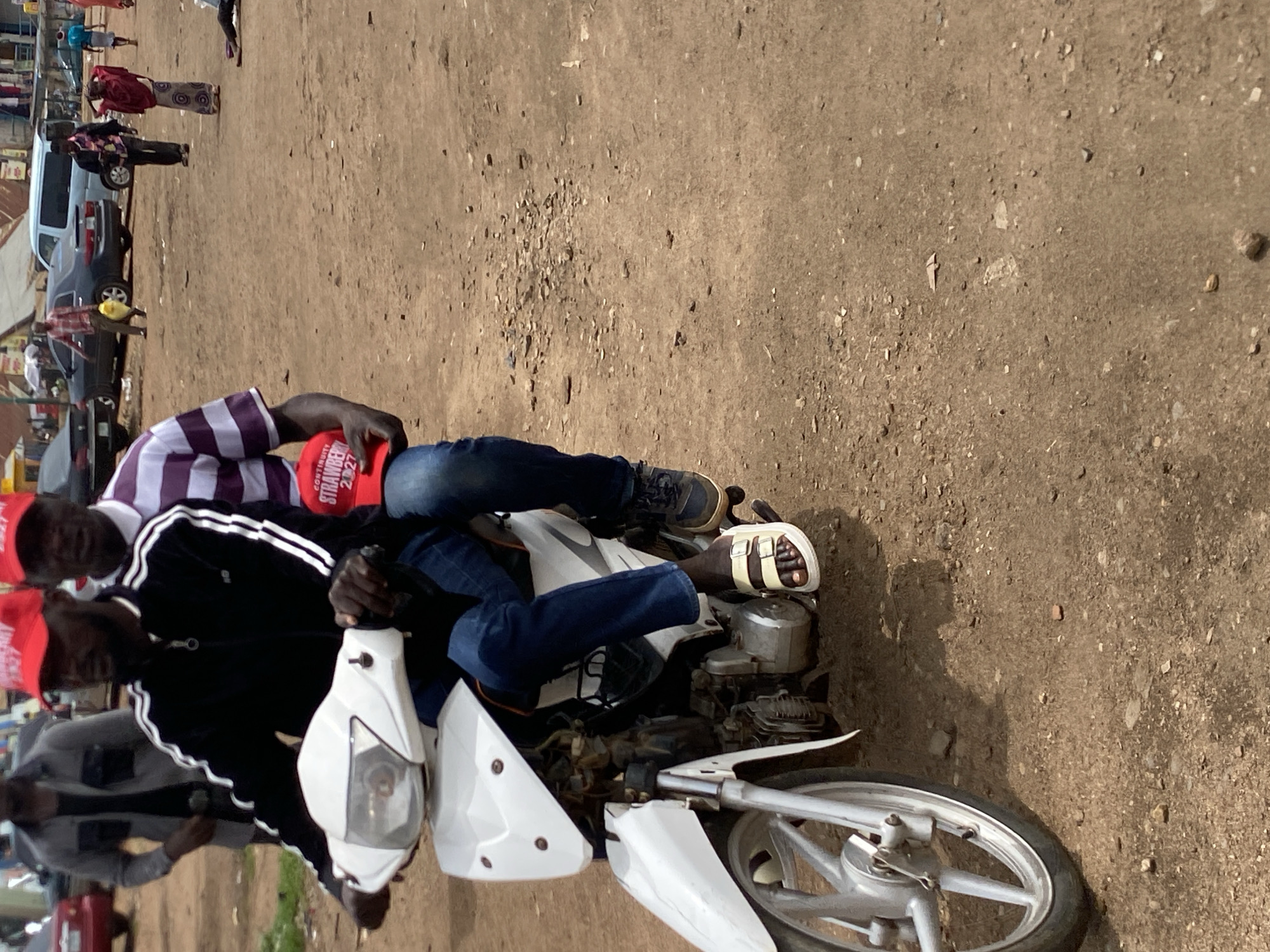 In Bukuru, Nigeria, two men are seated on a white motorcycle. The driver, facing forward, wears a black jacket with white stripes and a red cap. The passenger, sitting behind, wears a purple and white striped shirt, blue jeans, and a red cap. The setting appears to be a dirt road alongside a street with parked vehicles.