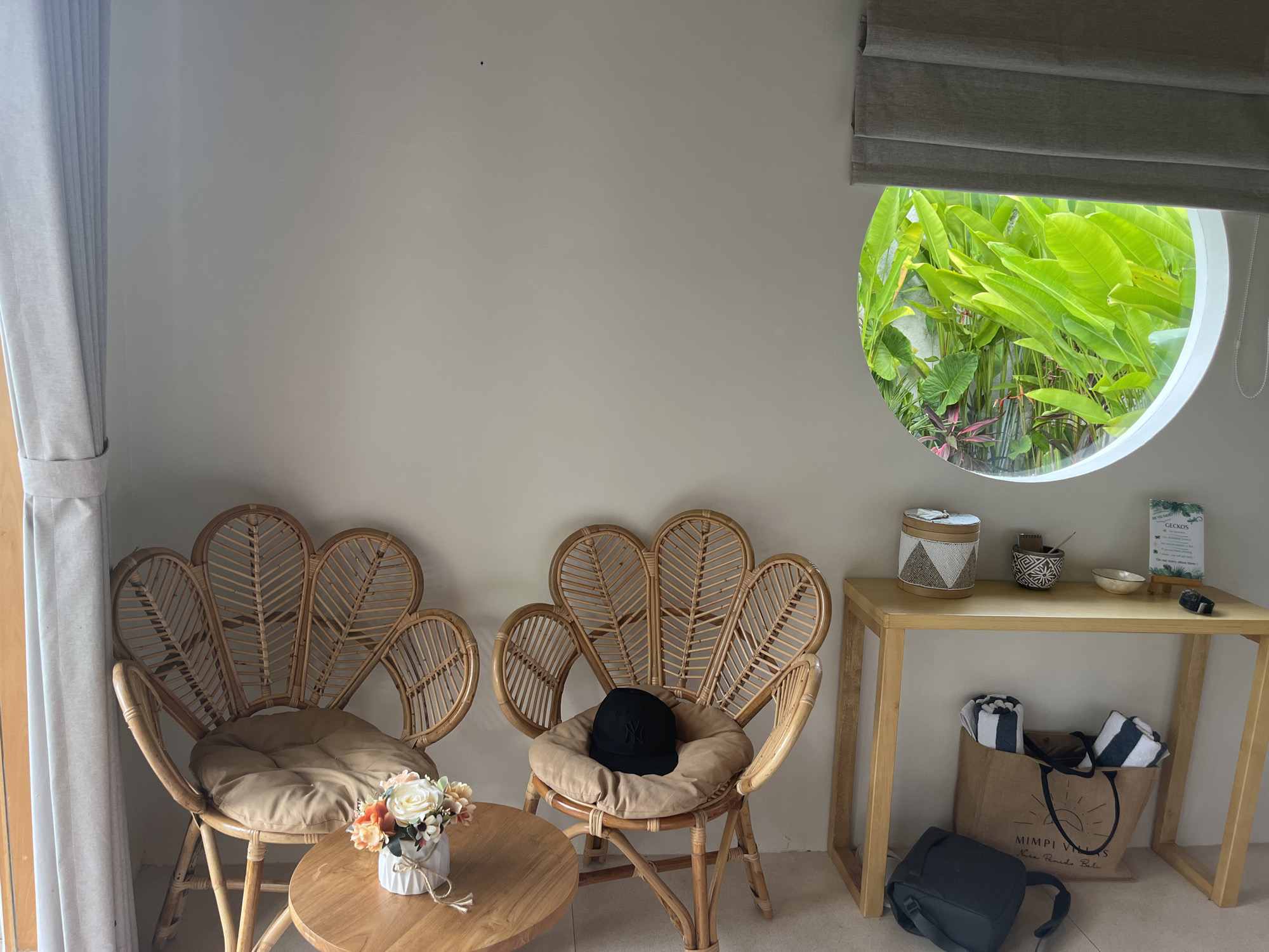 A daytime interior scene in Nusa Penida, Indonesia shows two rattan chairs with cushions, one with a black baseball cap on it. A small round wooden table sits between the chairs, holding a white pot of flowers. To the right, a light-colored wooden table holds a woven basket, a small bowl with implements, a small white bowl, a sign with the word "GECKOS" on it, and a small black object. Under the table, there is a tote bag with the words "MIMPI VILLAS Nusa Penida Bali" and a black camera bag. A round window with a gray shade above it shows green plants outside. A curtain is visible on the left.