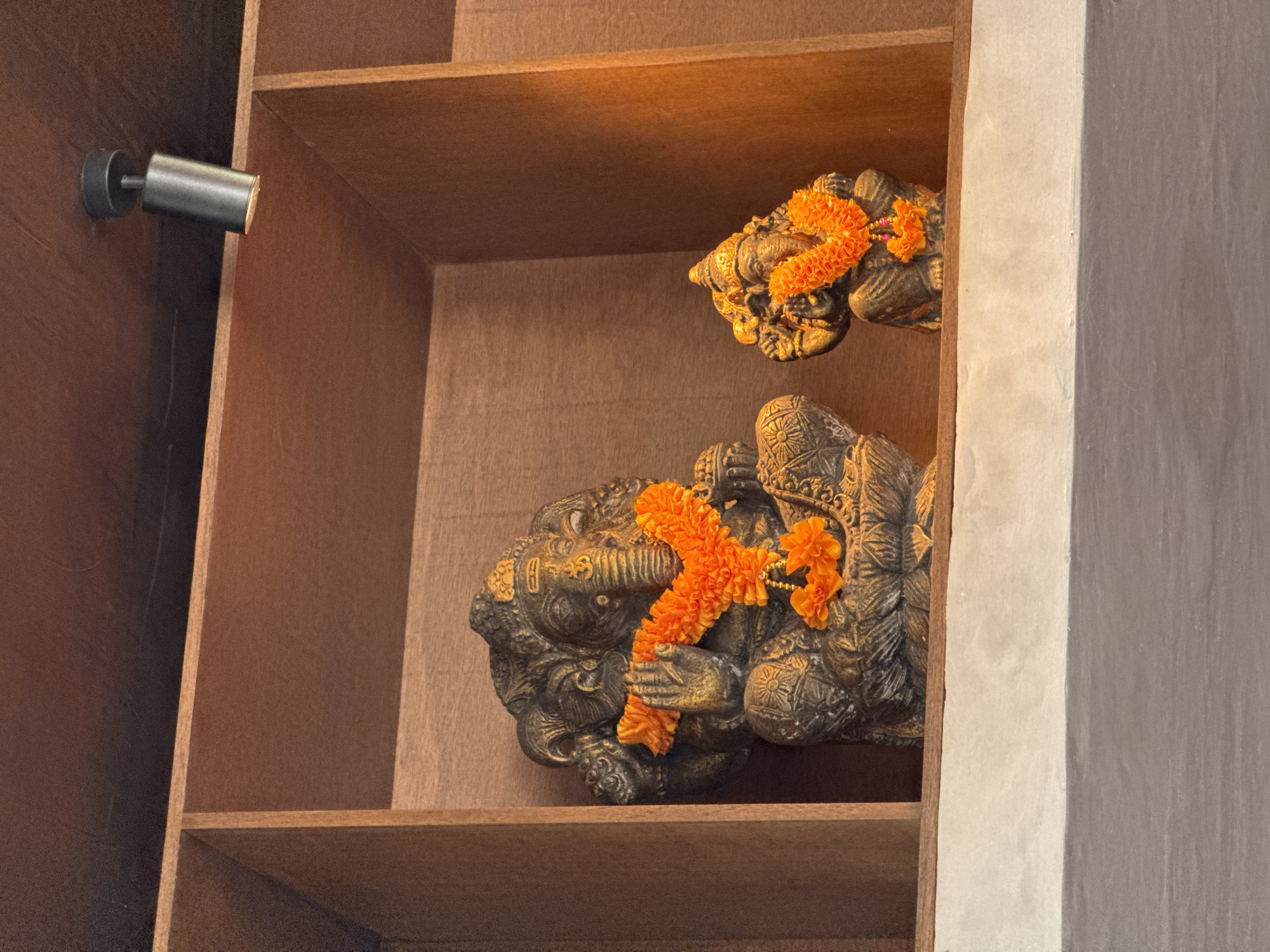 Two carved statues of Hindu deities adorned with orange garlands rest in a wooden shelf unit. A small spotlight is mounted on the unit's side. The location is Kuta Selatan, Indonesia.
