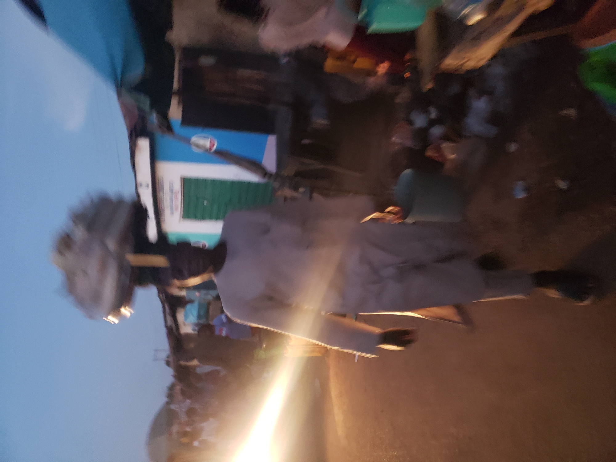 In Kakuri Gwari, Nigeria, at dusk, a person in a light-colored tunic and pants walks down a dirt path. They carry a large, round object balanced on their head, partially obscured by motion blur. To their right, a stall with a blue awning and a blue and white building with a green door are visible. Another person is partially visible to the far right, wearing a light-colored garment. The ground is a mix of dirt and debris, with a bright light source emanating from the left, casting a strong glow. The overall atmosphere suggests a bustling market or street scene as daylight fades.