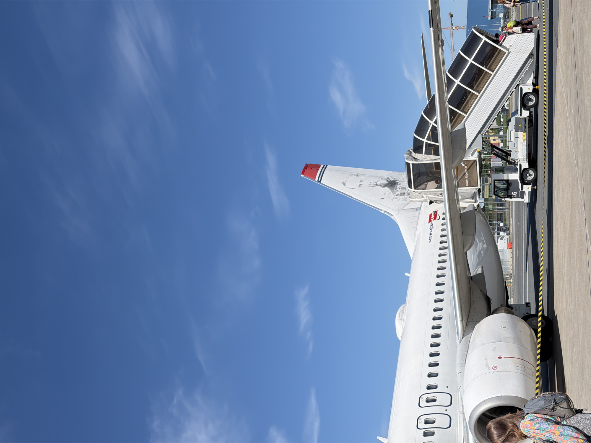 A Norwegian Airlines airplane with a portrait of Otto Sverdrup on its tail fin is parked at Tallinn Airport in Tallinn, Estonia. A mobile airstair, labeled "16", is connected to the plane's door, and passengers are boarding. A woman with a backpack stands on the tarmac near the plane's engine. The sky is clear and blue with a few wispy clouds. The airport tarmac is visible in the foreground, with a yellow and black striped safety line marking off the area. In the background, construction cranes are visible, suggesting ongoing development at the airport.