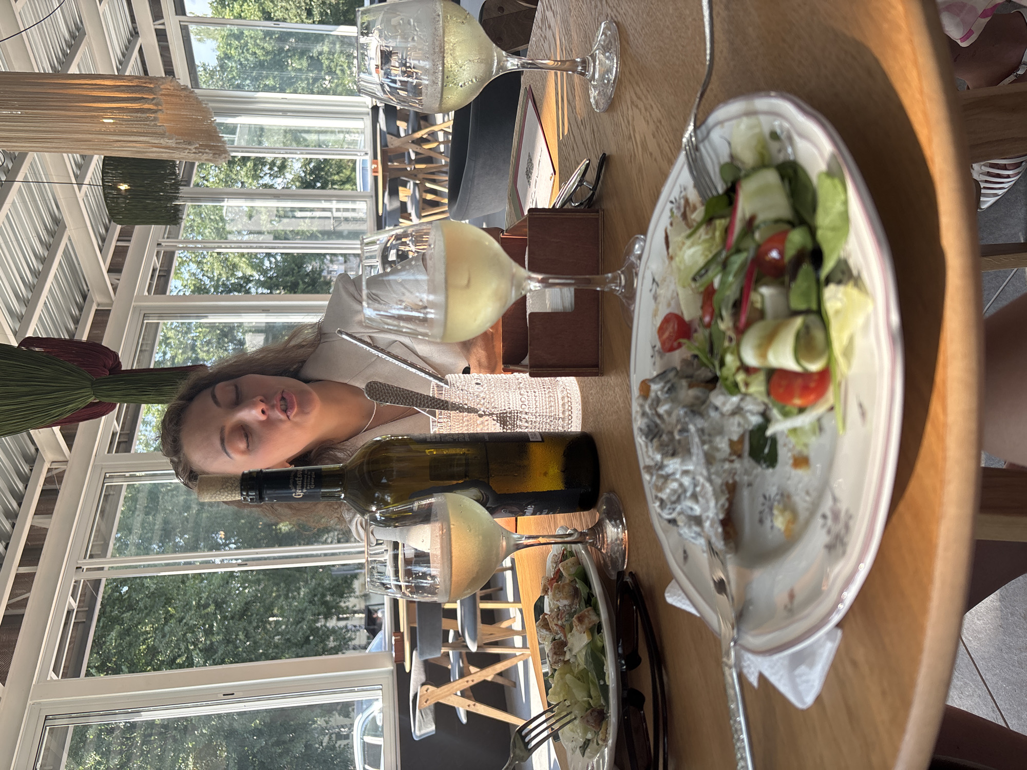An eye-level medium shot captures a woman at a table with food and drinks in a restaurant with large windows overlooking trees. The woman, with long brown hair, wears a white blazer and is talking with her mouth open. In front of her are two glasses of white wine, a bottle of white wine, a plate of food with a salad and what appears to be a mushroom dish, and another plate with a salad. The restaurant has a modern interior with hanging light fixtures and wooden furniture. The scene is brightly lit by natural light streaming through the windows. The setting is identified as Chaikovsky, Russia.