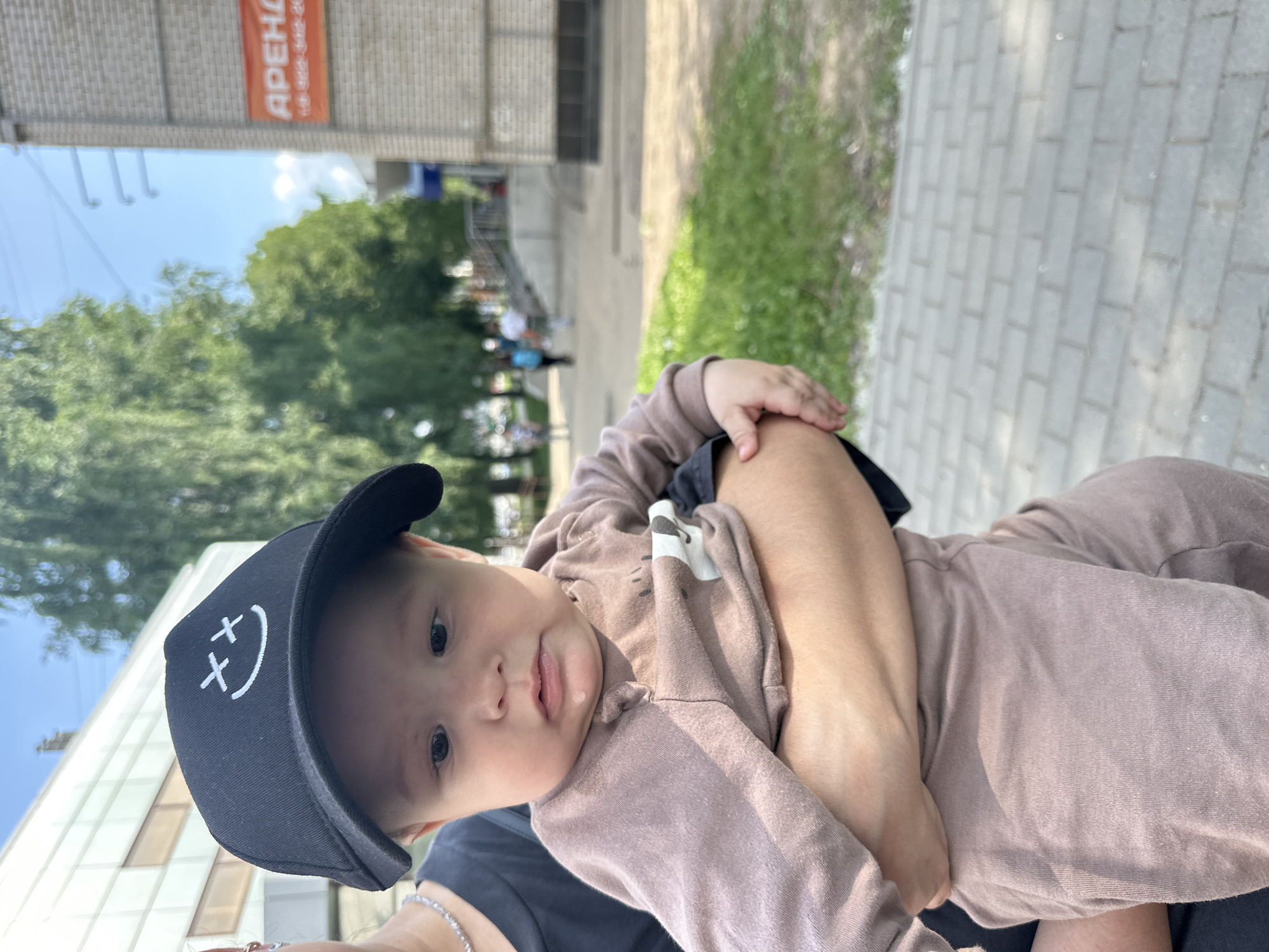 A close-up shot shows a baby wearing a black baseball cap with a white smiley face design. The baby is held in someone's arms, with their face looking down and slightly to the side. The baby is wearing a brown long-sleeved shirt. In the background, a brick building with an orange sign that reads "АРЕНДА" (rent) is visible. Trees and a clear blue sky are also present, suggesting an outdoor urban setting. The ground is paved with gray bricks.