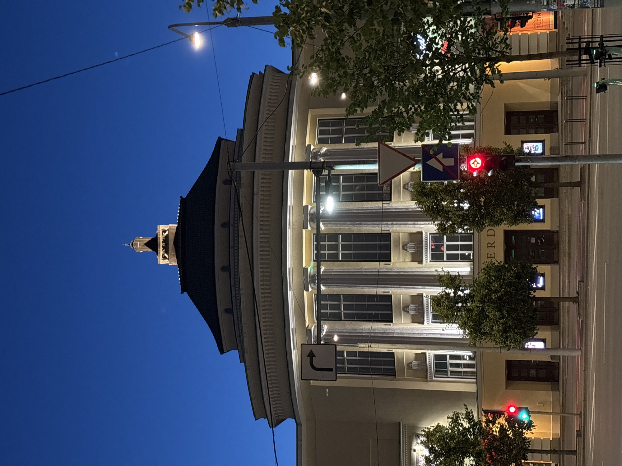 The image shows the exterior of a large, pale yellow building with classical architectural elements, illuminated at dusk. The building features a prominent circular base with tall windows, topped by a darker, curved roof and a central tower with a small cupola. Fluted columns are visible between the windows. A street with a traffic light showing red and green is in the foreground, with trees lining the sidewalk. The sky is a deep blue, indicating twilight. Street signs and utility lines are also present. Based on the architectural style and the text visible on posters on the right side of the building, which reads "FESTIVAL" in English and appears to be in Estonian, this building is likely the Estonia Theatre in Tallinn, Estonia.