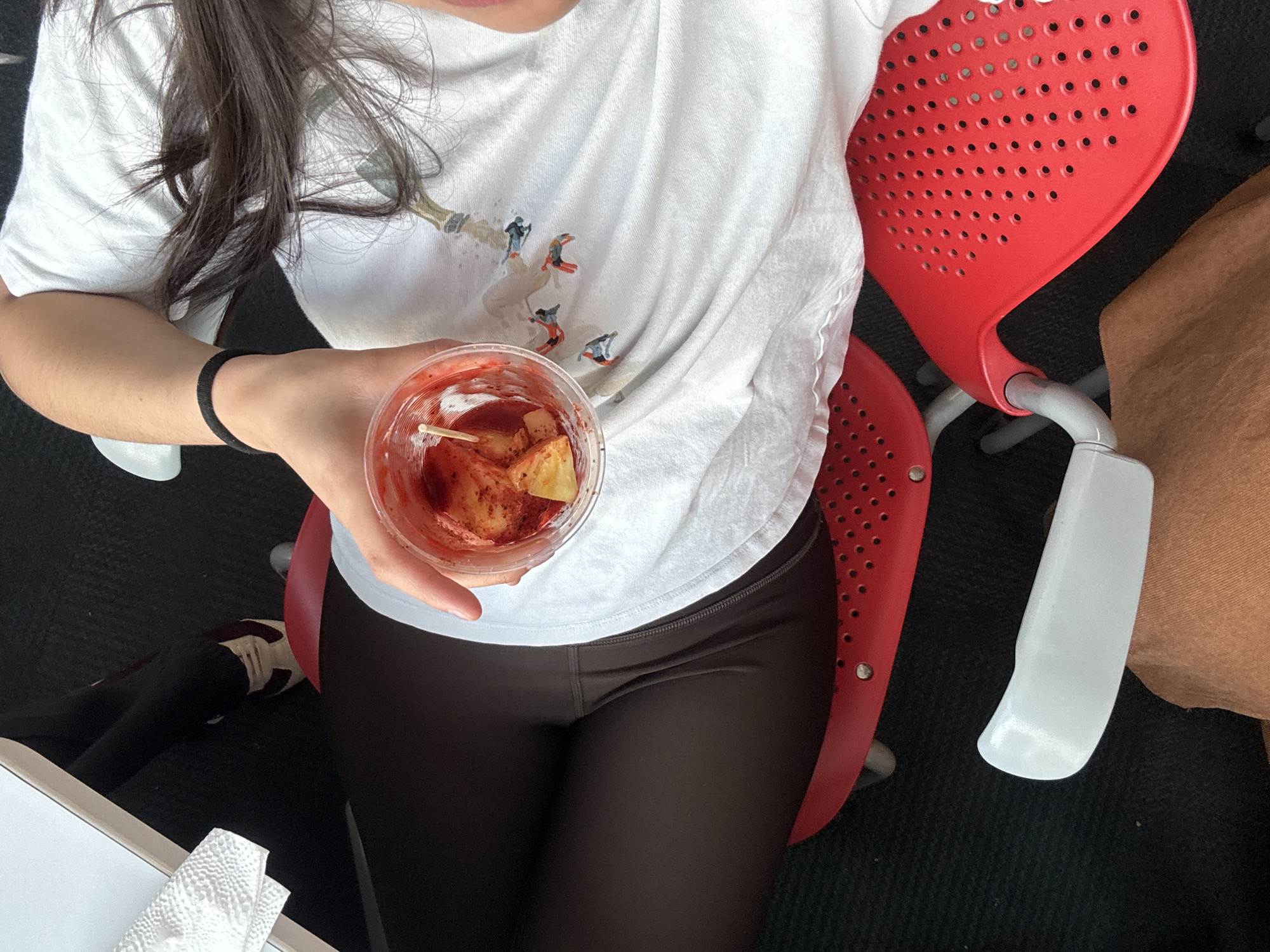 A top-down view depicts a person seated on a red chair. The person, whose head is out of frame, is wearing a white t-shirt with a design of skiers, and dark-colored leggings. In their hand, they hold a clear plastic cup containing what appears to be a red liquid and fruit pieces, possibly mango with chili powder. A black elastic band is visible on their wrist. To the left, there is a white surface and some folded white tissues or napkins. The red chair has a perforated design on the back and seat. To the right of the chair, a portion of a brown bag or surface is visible. The background appears to be a dark, possibly carpeted floor. The image provides no specific visual clues to suggest a location in Heroica Puebla de Zaragoza, Mexico.
