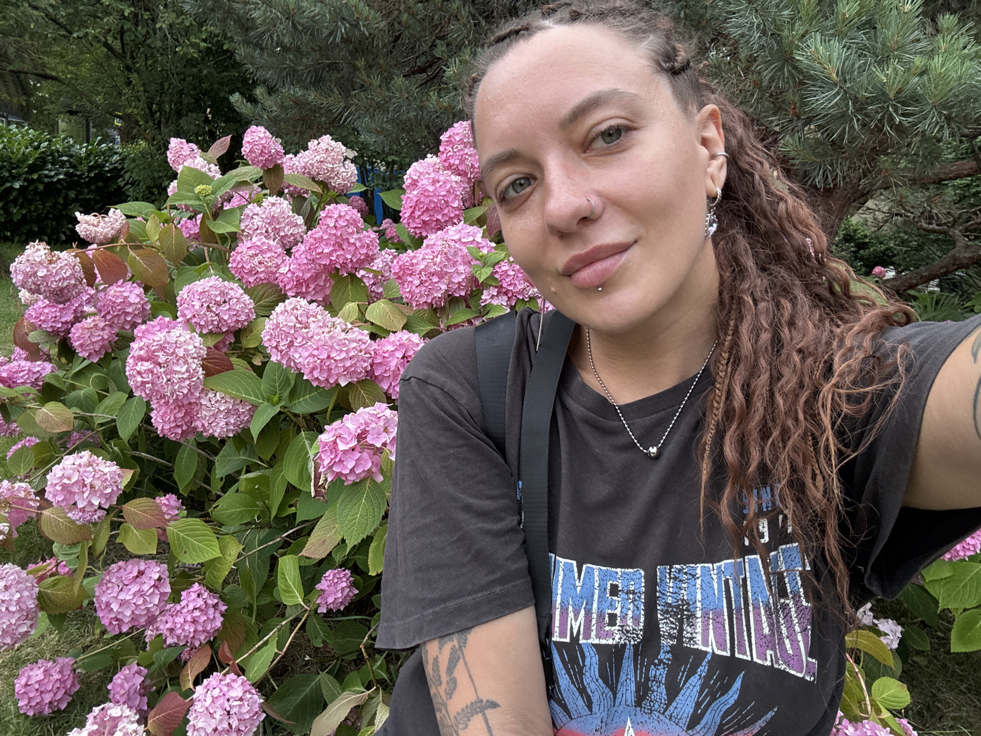 A medium shot photograph captures a woman with braided dark brown hair and piercings on her face, taking a selfie outdoors in front of a lush bush of pink hydrangeas. She is wearing a dark t-shirt with a graphic design. The background consists of green foliage and trees, suggesting a park or garden setting. The scene does not provide specific visual cues to identify the city as Sochi, Russia, nor does it offer any explicit indication of the current date or time.