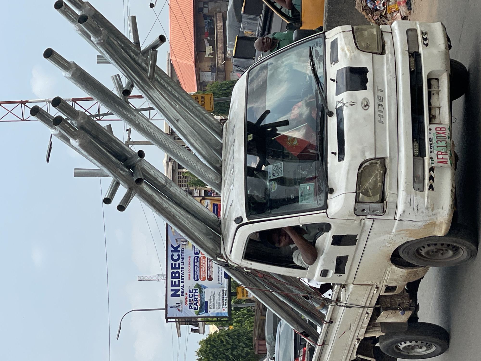 A white Daihatsu Hijet pickup truck, visibly dirty and with minor body damage including missing headlight surrounds and tape/paint on the hood, drives on an asphalt road in a city in Imo State, Nigeria. The truck is heavily loaded with at least four large, metallic, cylindrical structures, likely street light poles or similar infrastructure, secured with red straps. These poles extend over the truck's cab and significantly beyond its rear bed.
A dark-skinned male driver is visible in the cab, looking left with his arm resting on the open window frame. A small red flag is visible inside the cab on the dashboard. Another dark-skinned male in a green polo shirt stands on the right side of the truck, also looking left, near a yellow vehicle or object.
In the background, a large billboard for "NEBECK REAL ESTATE LIMITED" is prominent, advertising land for sale in various Nigerian locations, including "KINGS COURT ESTATE," "GREENVILLE ESTATE OBA PHASE 1AB," and "PURITY GARDEN ESTATE, ASABA." A black street light pole stands to the left of the billboard. Further back, a red and white lattice tower is visible under an overcast sky. To the right of the truck, a building with a reddish-orange corrugated metal roof features laundry hanging on a railing. A yellow street sign partially reads "NO LOADING" and "THE PARKS." A pile of refuse is visible on the far right.