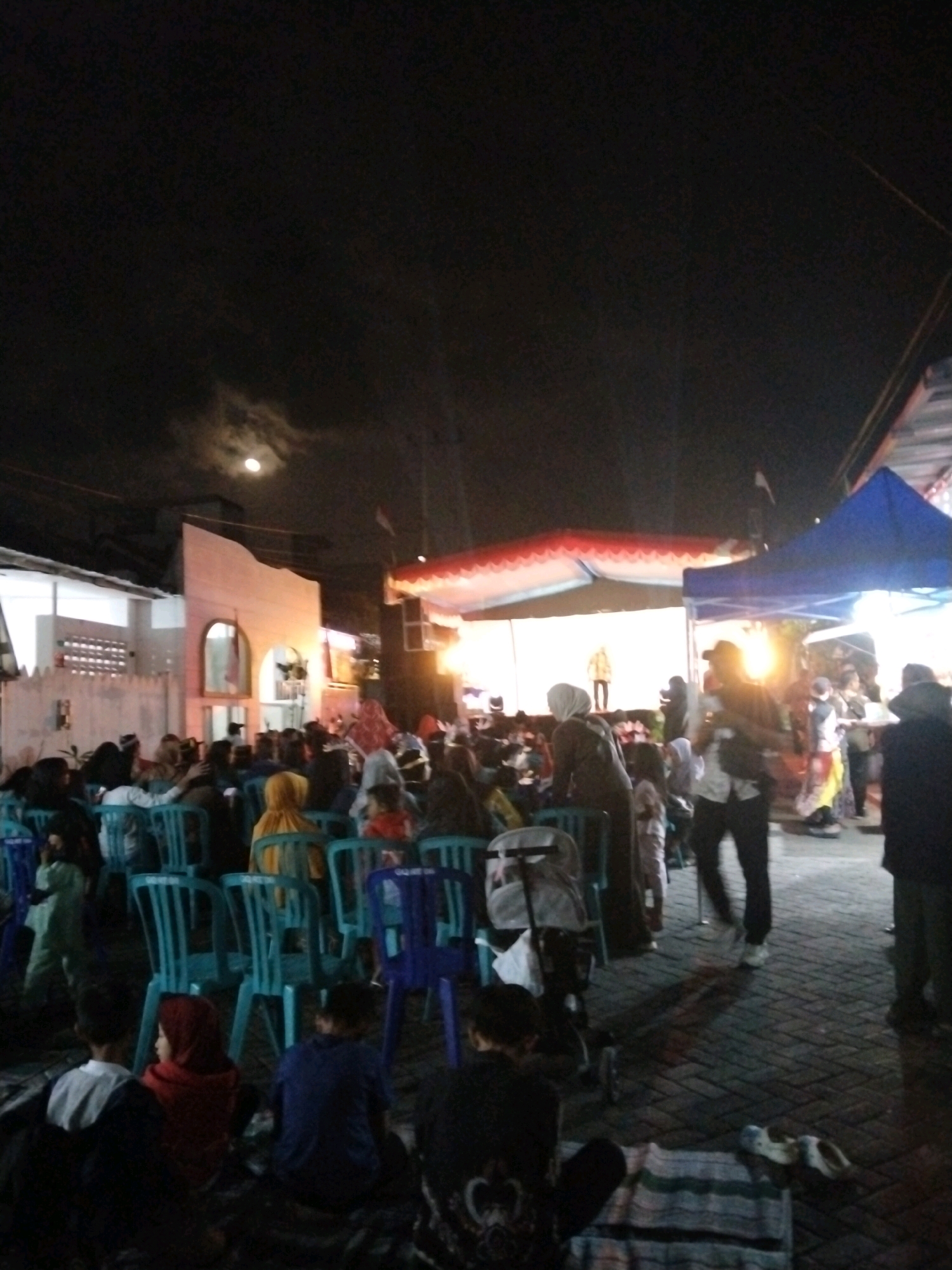 The image depicts an outdoor gathering at night in Singosari, Indonesia. The scene is illuminated by artificial lighting and moonlight, with a visible moon in the upper left portion of the sky. A crowd of people, many wearing hijabs, are seated on blue plastic chairs or on the ground, facing a stage or performance area. There are structures with awnings or tents set up, suggesting an event or market. Several individuals are standing, some appearing to be organizers or performers. Objects like a stroller and a mat are visible on the ground. The overall atmosphere is that of a community event taking place in a public space.
