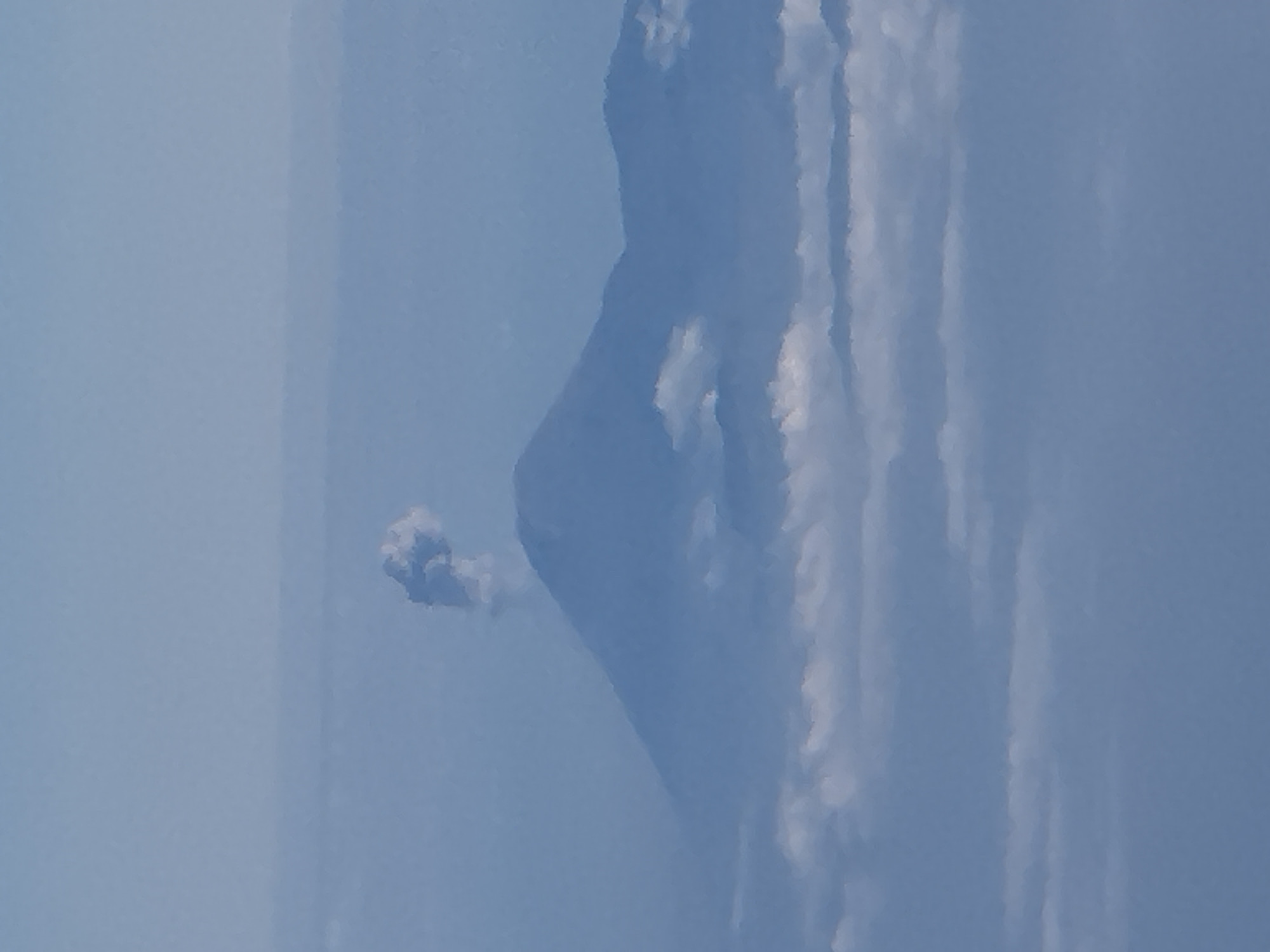 The image shows a volcanic eruption in a sky setting. A plume of ash and smoke billows from the peak of a volcano, which is partially obscured by clouds. The sky is a pale blue, suggesting it is daytime. The overall impression is one of raw natural power and the dynamic forces of the Earth. There is no visible text in the image. The scene depicts a moment of intense geological activity.