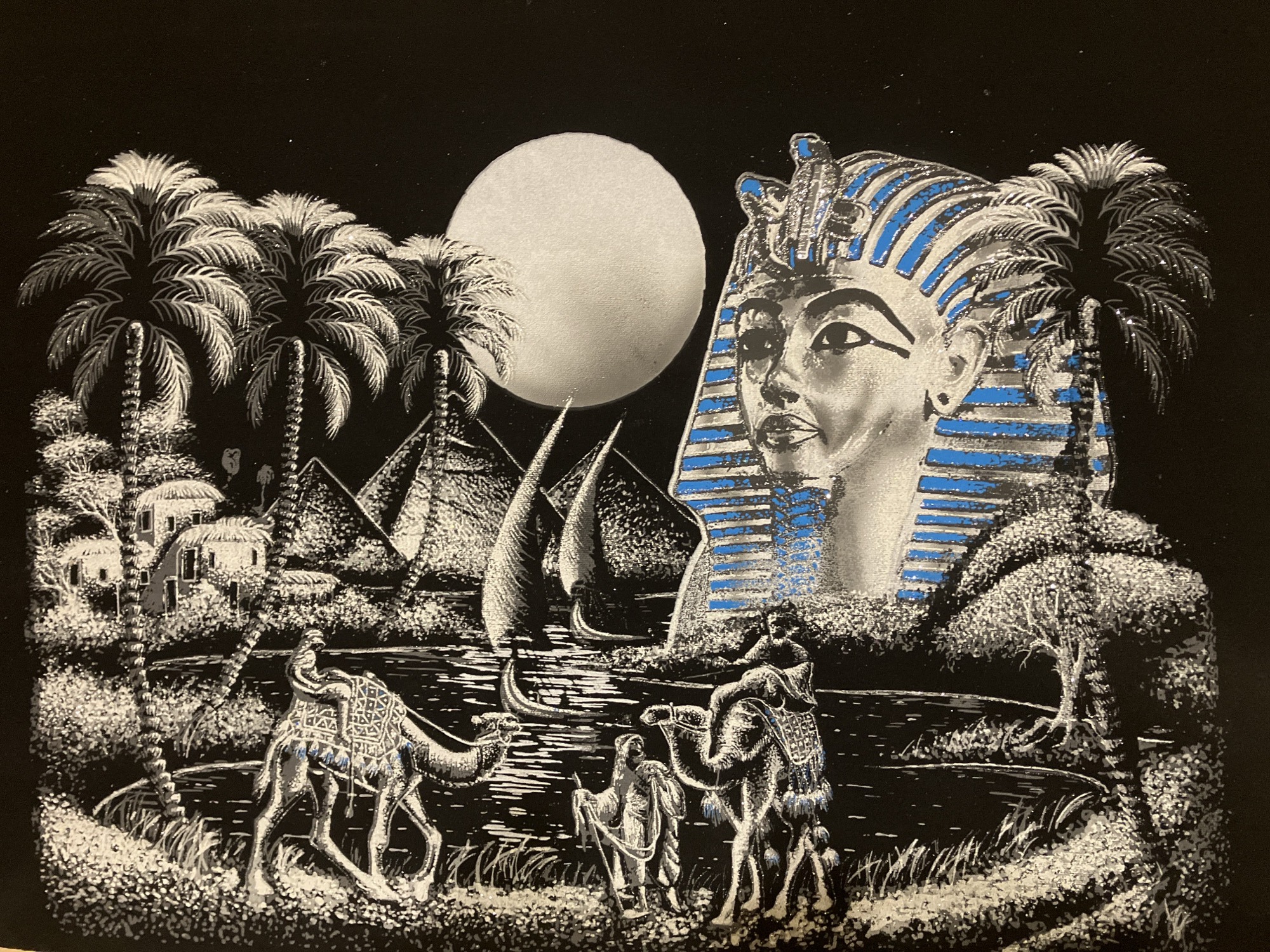This media file depicts an ornate, stylized scene set in ancient Egypt, rendered primarily in black, white, and silver with vibrant blue accents. The artwork appears to be a black velvet painting or scratchboard, characterized by a dark background with light, often glittery, elements.
In the upper right, a large, prominent head of a pharaoh, resembling Tutankhamun, dominates the scene. The pharaoh wears a striped nemes headdress, with alternating silver/white and bright blue bands, and a visible Uraeus cobra emblem on the forehead. Its gaze is directed forward.
Behind and to the left of the pharaoh's head, a large, luminous, circular orb (likely the moon) glows brightly in the dark sky. Several tall palm trees with intricate fronds are scattered across the composition, particularly on the left and right sides.
In the mid-ground, to the left of the moon, three triangular pyramids are visible, partially obscured by palm trees and other vegetation. A body of water, possibly the Nile River, flows through the mid-ground, reflecting the moonlight in shimmering streaks. Several traditional sailboats (feluccas) with triangular sails navigate the water.
The foreground features a riverbank lush with vegetation, depicted in white and silver. A caravan of camels and people traverses this area. On the left, a camel with a decorative saddle and a rider walks along the bank. In the center, two figures, one with a staff, walk alongside another camel, also adorned with intricate saddle blankets featuring blue details.
Further in the background on the left, nestled among the palm trees, are several small, round-roofed dwellings or huts, adding to the depiction of an ancient Egyptian settlement. The overall scene evokes a sense of exotic mystery and ancient grandeur under a moonlit sky. There is no visible text in the image.
