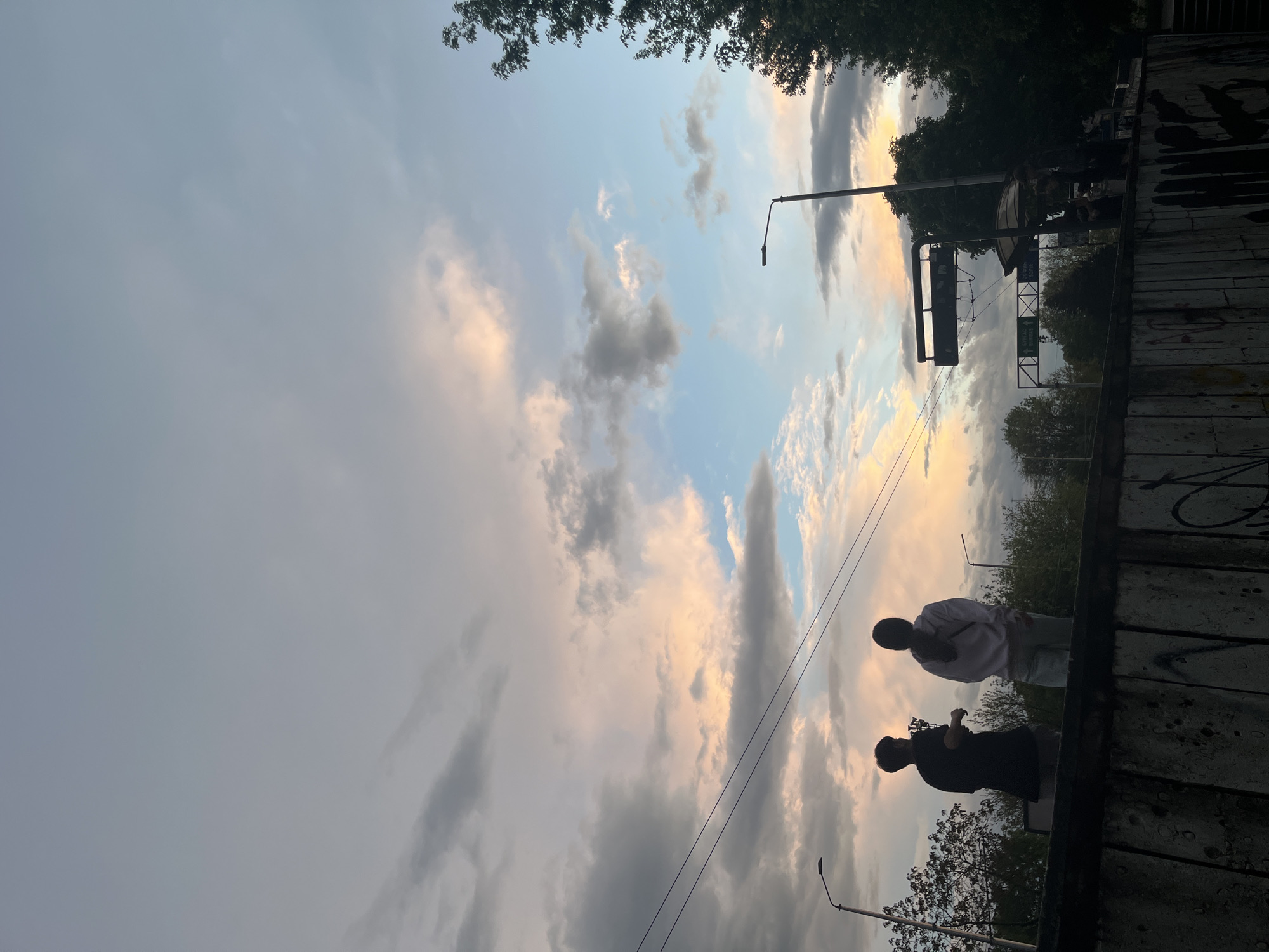 The image shows two silhouetted figures standing on a concrete structure, likely a bridge or elevated walkway, against a backdrop of a sky with scattered clouds and patches of blue. The sky exhibits colors suggesting either sunrise or sunset. Street lamps are visible in the frame. In the upper portion of the image, the silhouettes of trees obscure portions of the sky. The scene appears to be an urban environment. The location is Varna, Bulgaria. The image contains a traffic sign structure. Two individuals are discernible on the structure, one seemingly holding a phone or camera, potentially engaged in photography. They are interacting with one another.