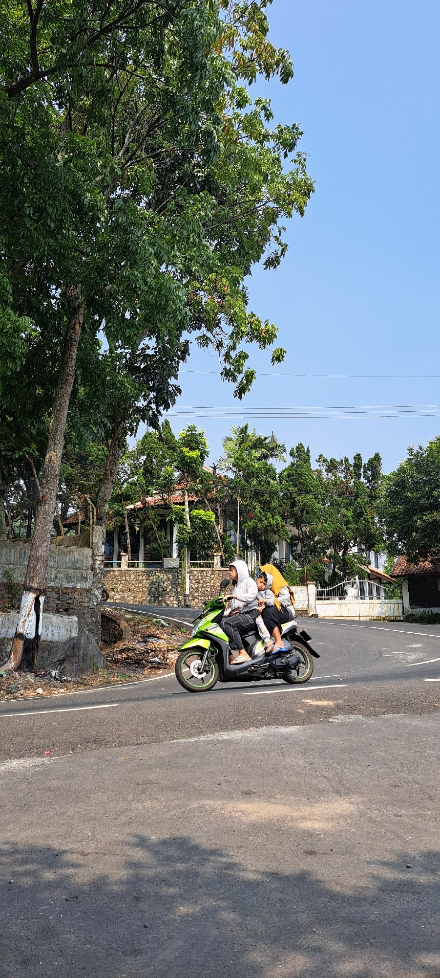 The image captures a street scene in Prigen, Indonesia, on a clear day. Three individuals are riding on a lime green scooter. The person in the front, wearing a grey hooded jacket and black pants, appears to be driving. Seated behind the driver is a child wearing a grey outfit. A third person, wearing a yellow headscarf and a grey top, is the rear passenger. The scooter is on an asphalt road with white lane markings. To the left of the road is a textured concrete barrier with some areas painted white and black, next to a large tree with lush green foliage. In the background, several buildings with tiled roofs are visible, surrounded by various types of trees, including palm trees. Power lines are strung across the sky, which is a bright blue. Shadows of trees are cast on the road in the foreground.