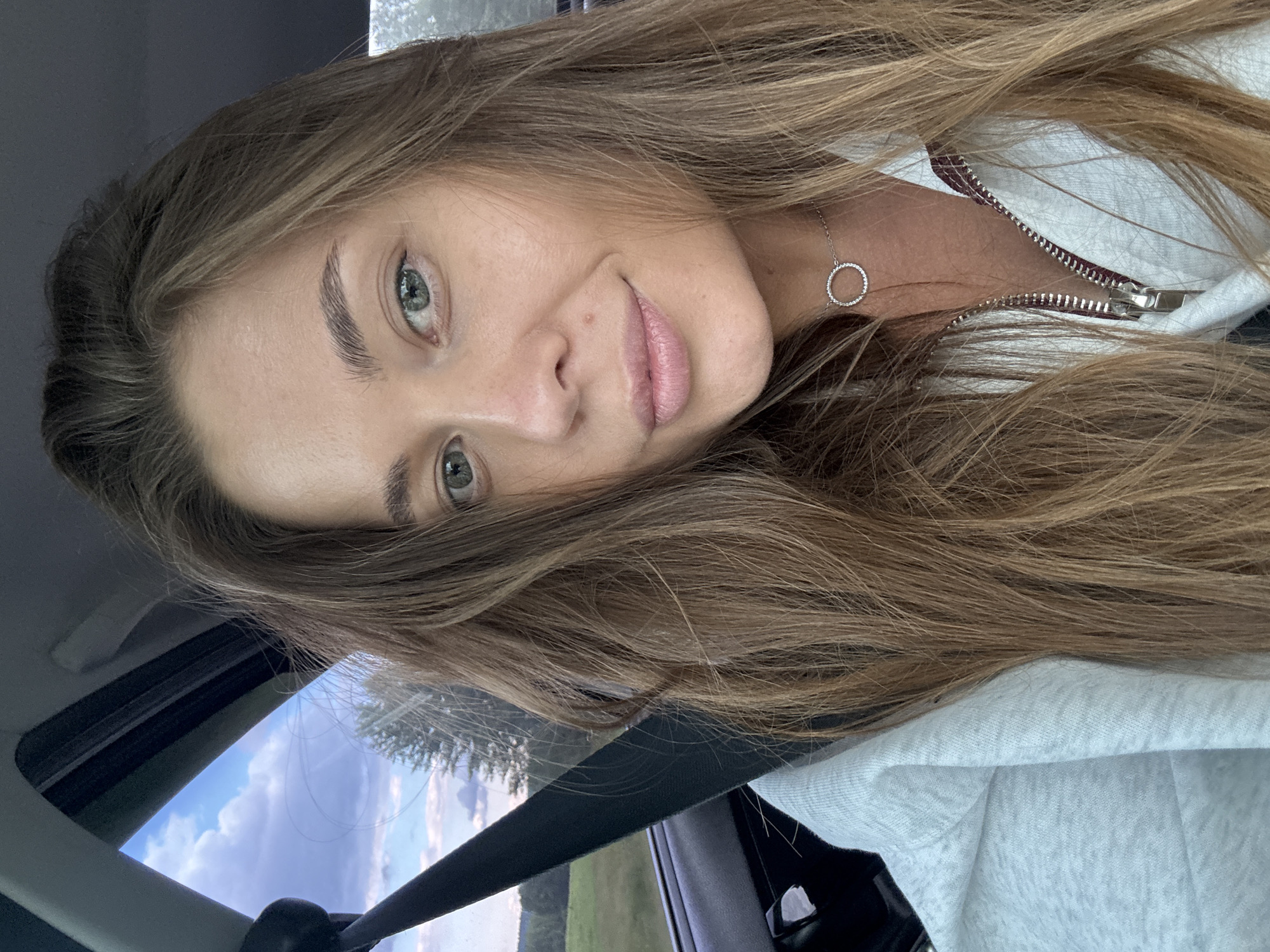 A close-up, selfie-style image shows a woman with long, light brown hair and green eyes. She is sitting in the passenger seat of a car, indicated by the visible seatbelt and car interior. The woman is wearing a light-colored, zippered hoodie and a delicate necklace with a circular pendant. The background outside the car window shows a landscape with trees and a cloudy sky, suggesting a rural or suburban setting, possibly on the outskirts of Бикбарда, Russia. The lighting is natural, suggesting it's daytime.