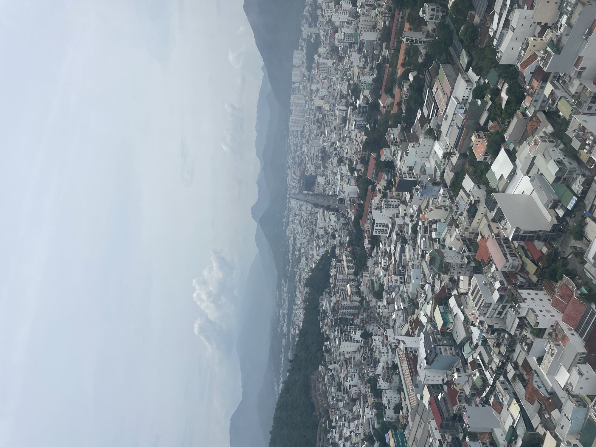 This is an aerial view of a dense urban landscape in Nha Trang, Vietnam. The foreground is dominated by a sprawling collection of buildings with a variety of architectural styles and roof colors, including white, grey, red, and green. Many structures appear to be multi-story residential or commercial buildings, interspersed with smaller houses and what seem to be rooftop solar panels on some of them. Lush green trees are visible throughout the city, peeking out from between buildings and along the hillsides.
In the mid-ground, a wide, straight road or highway cuts through the city, lined with taller, more modern buildings, including several high-rise apartment blocks. The urban sprawl continues further into the distance, meeting a backdrop of rolling green hills. The hills are densely covered with vegetation, and in the far distance, a range of larger, hazy mountains forms the horizon. The sky is overcast with soft, diffused light, with a few scattered white clouds. The overall impression is a coastal city with a significant population density, nestled between mountains and likely near the sea, although the sea itself is not visible in this frame. There are no visible people or vehicles that are distinct enough to be described.