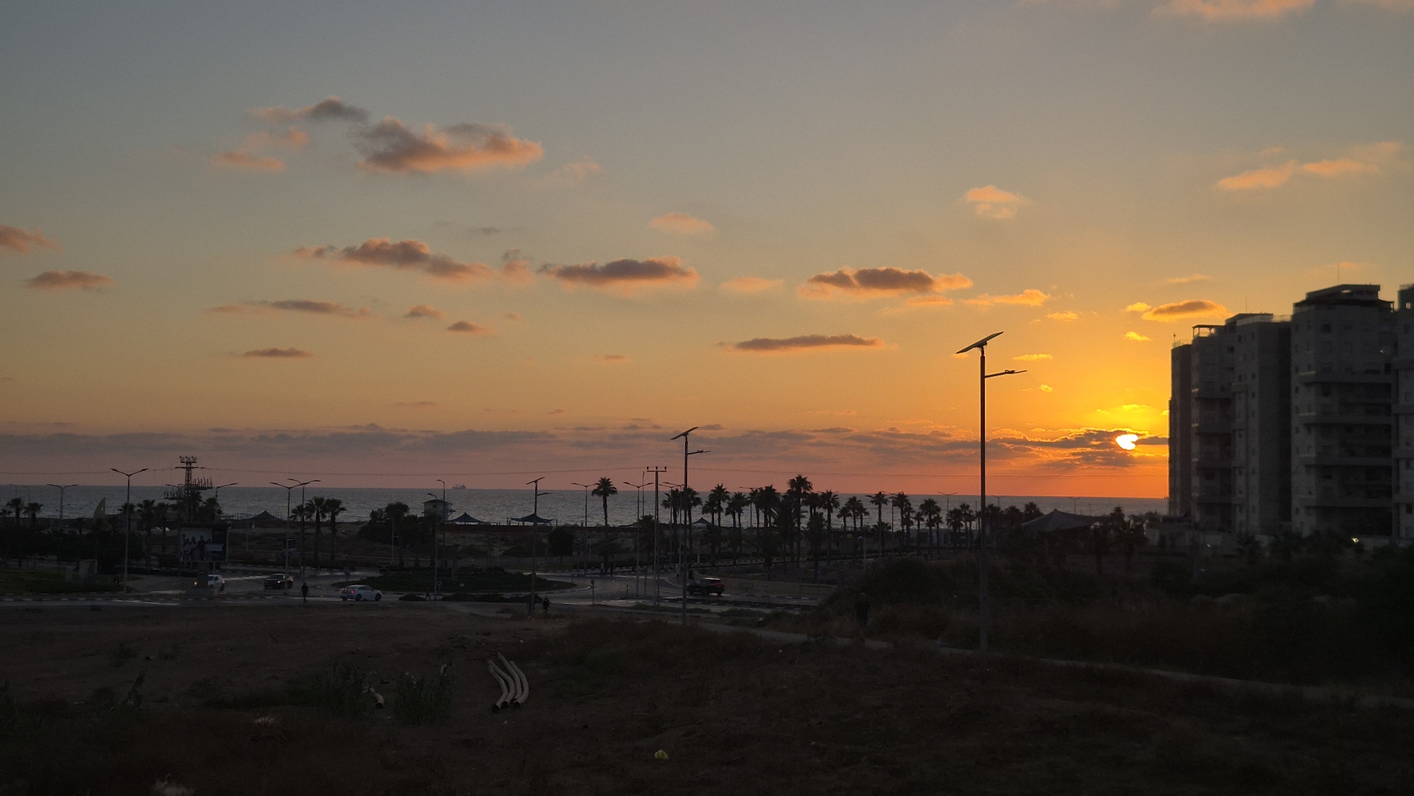 The image captures a sunset over the city of Ashdod, Israel. The sky is a gradient of orange and pink, with scattered clouds illuminated by the setting sun. In the foreground, there is a dark, silhouetted landscape with some vegetation and debris.
In the middle ground, a road runs horizontally across the scene, with a few cars visible. Palm trees line the road, and beyond them is the sea. There are several tall lampposts with solar panels along the road. In the distance, a tall structure that appears to be a crane or tower is visible on the left side of the image.
On the right side of the image, several modern, multi-story buildings are silhouetted against the bright sky. The sun is partially obscured by clouds, casting a warm glow over the entire scene. The overall impression is one of a coastal urban environment at dusk.