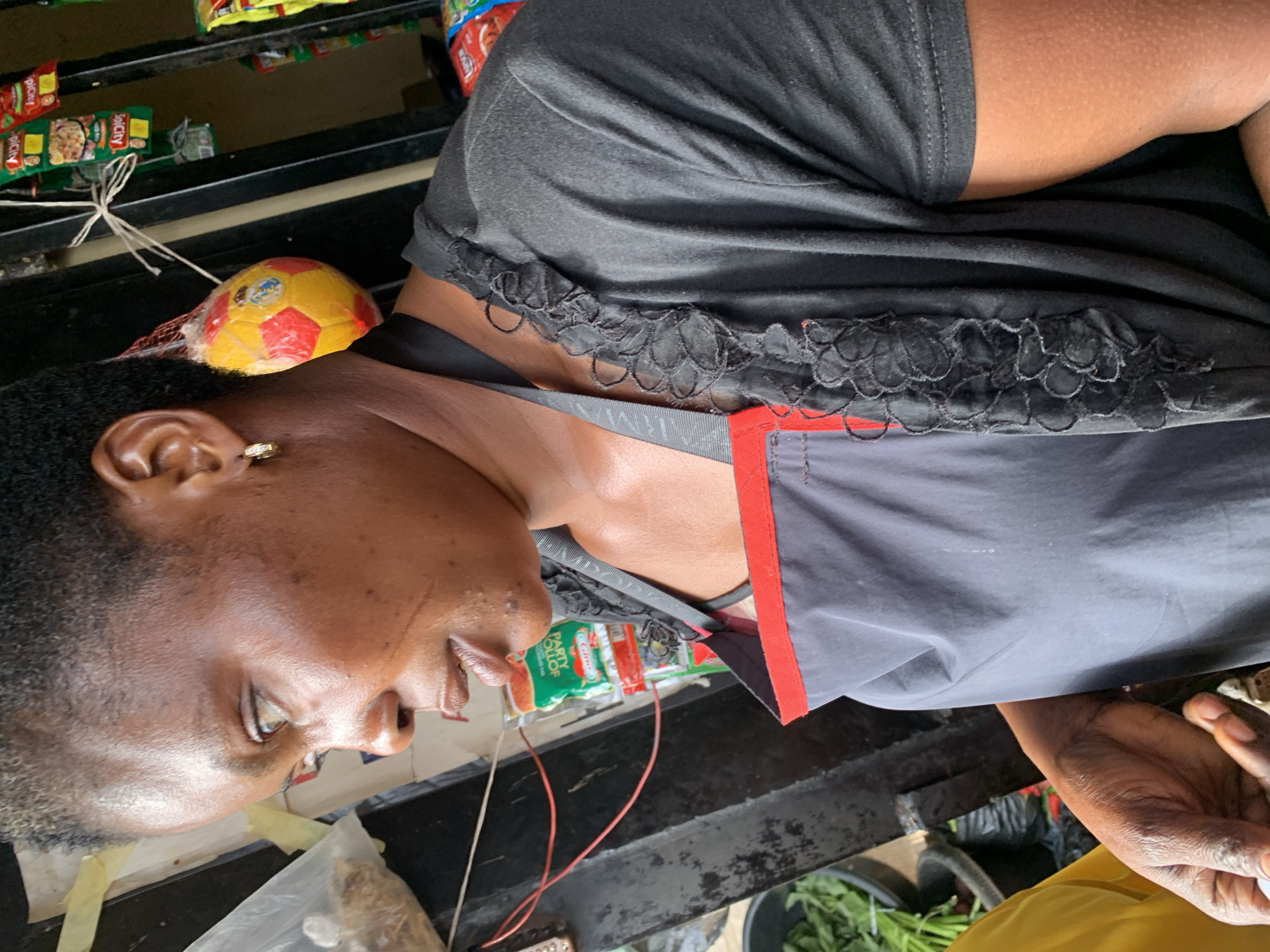 In Barakin Akawo, Nigeria, a woman is captured in a close-up shot, her profile facing left. She wears a black top and a grey apron with a red trim. A small, multi-colored toy ball, possibly a football, is suspended by a string from her hair, resting on her shoulder. Behind her, shelves are stocked with various packaged goods, suggesting a market or store setting. In the lower left corner, a black basin filled with green leafy vegetables is visible, along with a portion of a yellow garment belonging to another person. The lighting suggests an outdoor or well-lit indoor environment.