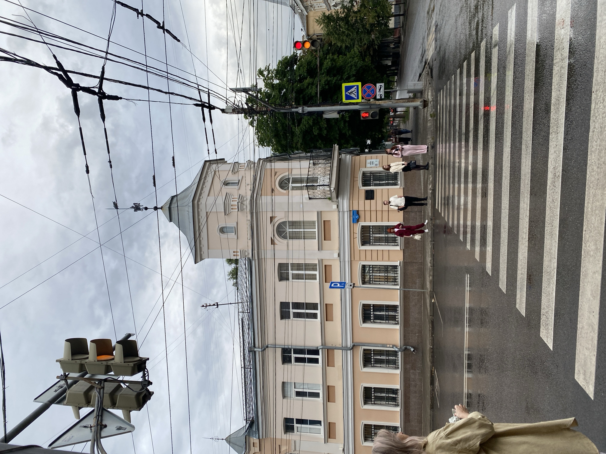 A street scene in Voronezh, Russia, depicts a pedestrian crossing under a cloudy sky. The crossing is wet, indicating recent rain. On the left, a woman with blonde hair, wearing a light yellow shirt, is visible from the side, holding a phone. Several people are waiting at the crosswalk on the sidewalk in front of a light pink building. A traffic light ahead shows yellow and red. Overhead, a complex network of electrical wires crisscrosses the sky. The building features ornate architectural details, including arched windows and a small balcony. In the background, more buildings and trees line the street, and additional traffic lights and street signs are visible.