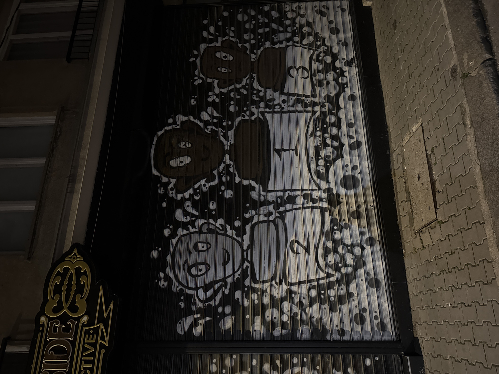 This nighttime image presents a vibrant urban scene, likely an alley or side street in Sofia, Bulgaria, dominated by a large, corrugated metal surface covered in graffiti. The artwork, set against a dark background, features cartoonish, blob-like characters and numbers rendered in white, light grey, and brown/dark grey.
The graffiti includes several figures with prominent eyes; two brown, amorphous shapes are visible at the top left, while a white-outlined, circular character with a smiling mouth appears at the bottom left. Bold numbers are integrated into the design: a "3" at the top right, a "1" in the middle, and a "2" at the bottom right, all within blocky, white-bordered shapes. The entire piece is enlivened by numerous white and dark grey circles resembling bubbles or paint splatters, creating a dynamic texture.
To the far left, an ornate black and gold sign is partially visible, displaying the text "IDE" and "CTIVE," suggesting part of a word such as "DETECTIVE." Above the sign, a section of a building facade with a window and a dark railing can be seen. On the right, the ground is paved with interlocking concrete blocks, typical of a sidewalk, alongside a curb and a stretch of rougher ground. The scene is illuminated by artificial light, confirming the nighttime setting. The overall mood is quiet, showcasing a blend of urban architecture and playful street art.