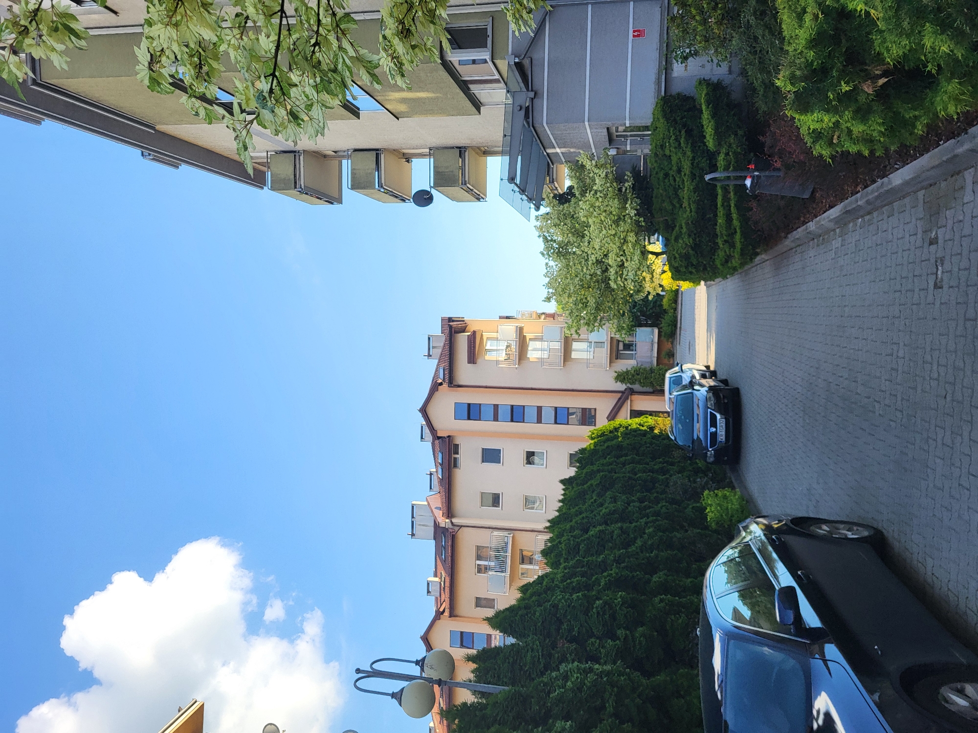 The image shows a street scene in Unknown. The street is paved with grey bricks. Two buildings flank the street: a light grey building with multiple balconies on the left, and a tan building, also with balconies, on the right. Trees and bushes line the sides of the street. Four cars are parked on the street. The sky is blue with scattered white clouds. A streetlight is visible in the bottom left corner of the image.