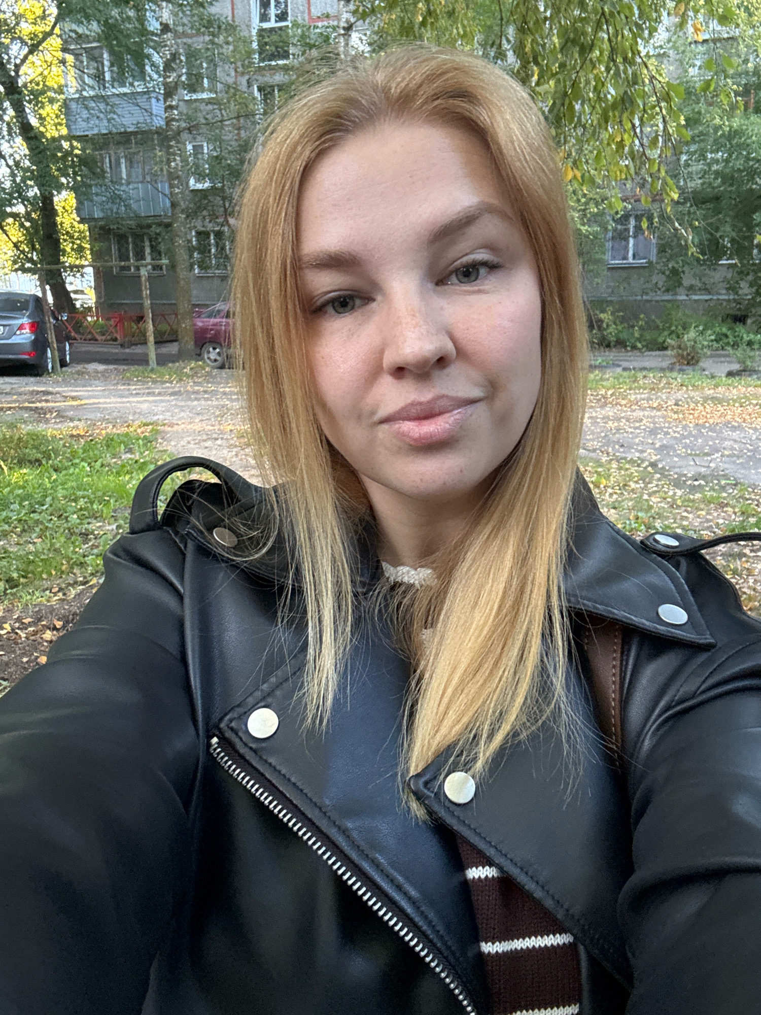 A young woman with blonde hair and light-colored eyes takes a selfie outdoors. She is wearing a black leather jacket over a brown and white striped shirt. In the background, there is a multi-story building, trees with green and yellow leaves, parked cars, and a paved area covered with fallen leaves, suggesting an urban residential setting in Rusanovo, Russia during autumn.