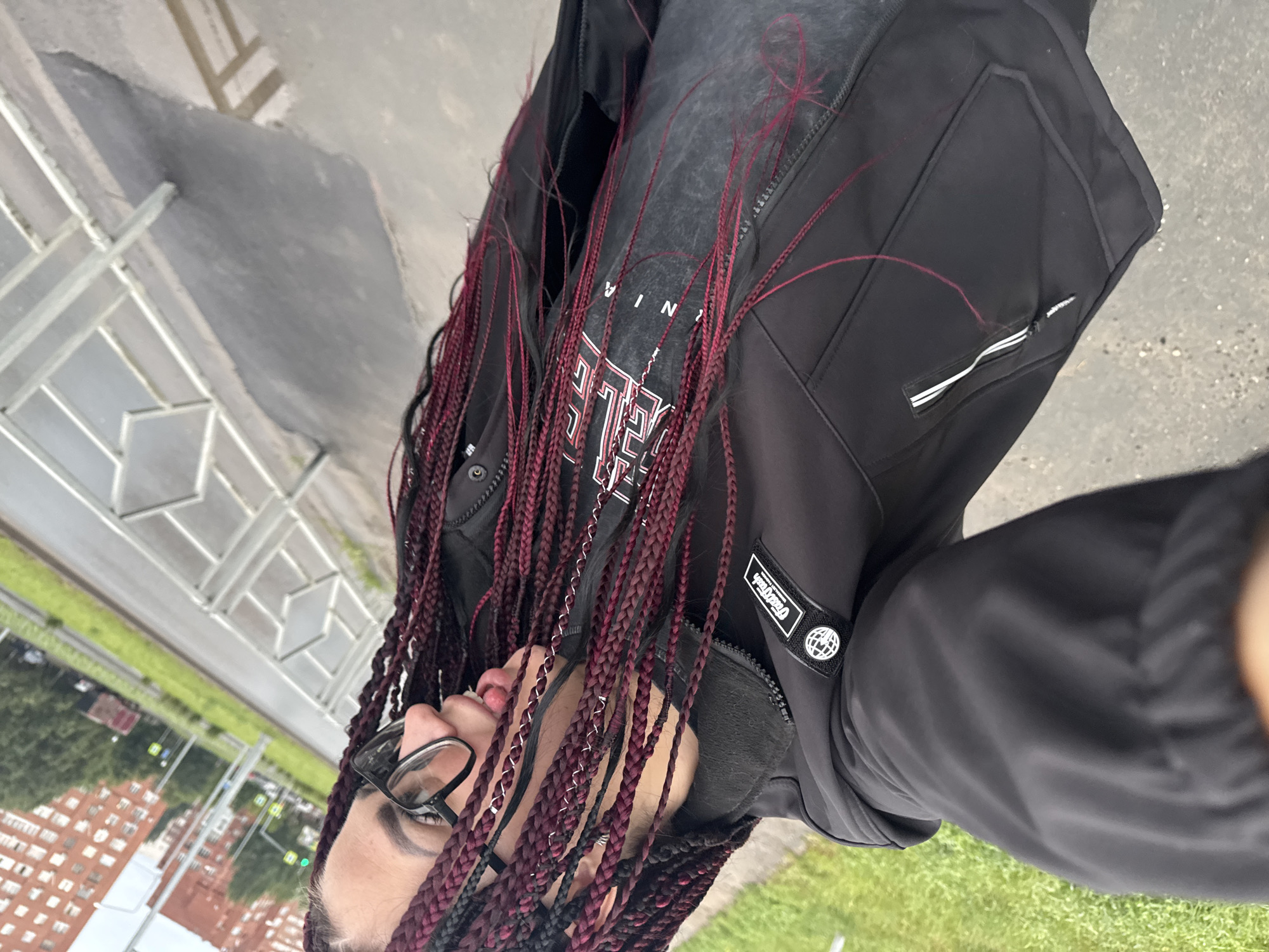 A young woman with long, braided hair, predominantly burgundy with some black strands, is taking a selfie. She is wearing black-rimmed glasses and a black jacket over a dark grey t-shirt with white text. Her tongue is slightly sticking out. She appears to be standing outdoors on a paved surface, possibly a sidewalk or walkway, next to a grassy area. In the background, a multistory brick building and a street with a metal railing are visible, suggesting an urban environment. The image is taken from a slightly tilted, upward angle, likely a street in Tolyatti, Russia, given the context.