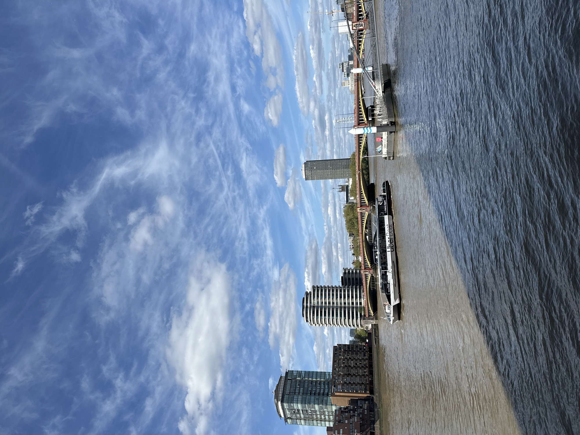 The image shows the River Thames in London, United Kingdom. In the foreground is a boat labeled "Uber Boat" docked at a pier. The river reflects the sunlight, creating a bright and shimmering surface. The opposite bank features modern buildings, including a cylindrical, multi-story structure with horizontal banding. Further down the river, a bridge is visible, constructed with a red and white steel frame. In the background, a tall, slim skyscraper rises above the other buildings. The sky above is blue, filled with scattered, wispy clouds.