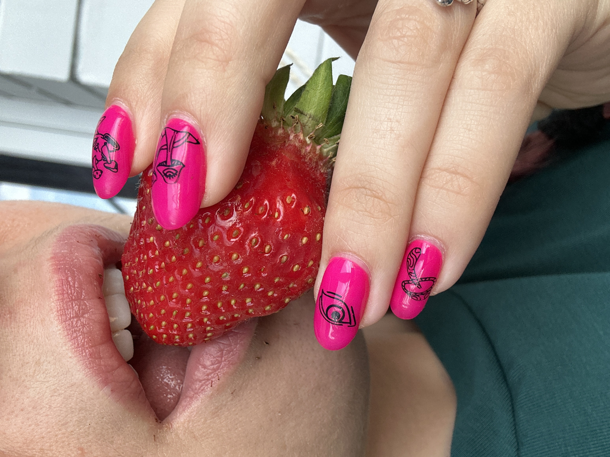 A close-up, brightly lit image captures a person's hands and mouth interacting with a large, ripe strawberry. The person, whose lower face, lips, and teeth are visible, appears to be holding the strawberry near their mouth, poised to take a bite. Their hands are manicured with vibrant hot pink nail polish, each visible nail featuring a distinct black line art design, primarily stylized eyes or abstract faces incorporating an eye motif. A silver ring with a subtle design is worn on one finger.
The strawberry itself is bright red with visible achenes (seeds) and its green calyx (stem and leaves) still attached. A dark teal or green fabric, likely clothing, is visible on the right side of the frame. The background is softly blurred, showing hints of a bright, light-colored structure, possibly a window frame or outdoor element. The bright lighting suggests it is daytime. The scene evokes a sense of freshness and enjoyment, set in Vladimir, Russia. There is no visible text in the image.