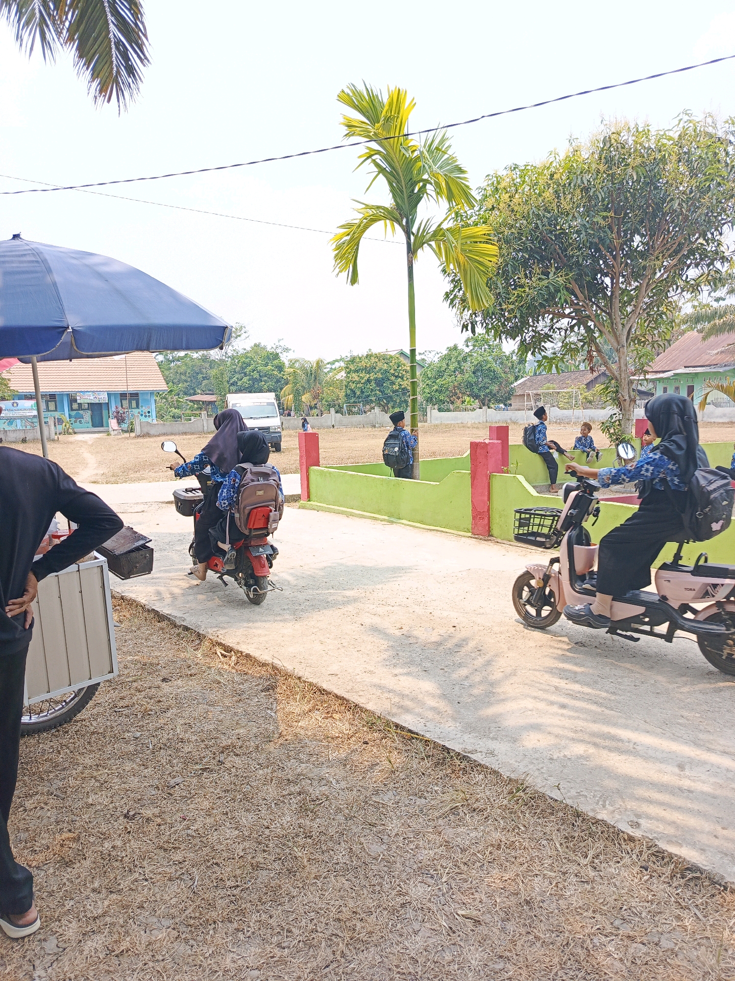 In Megang Sakti V, Indonesia, on a sunny day, a group of students in uniforms are seen in the vicinity of a school. The scene is characterized by a light-colored pathway, dry, yellowish grass, and a green-painted concrete barrier. In the foreground, a person dressed in black stands near a cart with a blue umbrella. Further back, two students, a female driver and a passenger, are on a red motorbike, both wearing hijabs and carrying backpacks. Across the pathway, a pink electric scooter is ridden by a student in a hijab, also with a backpack. Several other students in similar uniforms are visible in the background, some sitting on the green barrier and others standing or walking near a white van. A two-story school building with blue walls and a brown roof is situated behind a fence. Palm trees and other lush green foliage are present throughout the scene, suggesting a tropical environment. The overall impression is of a school dismissal or a similar activity involving students.