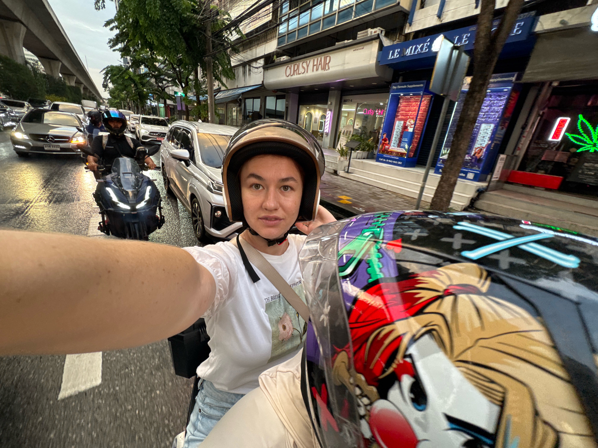 This is a selfie taken from the perspective of a passenger on a motorcycle, in Bangkok, Thailand. The passenger, a woman wearing a white t-shirt with a floral design and light blue jeans, is wearing a helmet. In the foreground, the passenger's arm and the helmet of the person in front of her, decorated with a cartoon character, are visible. The rider in front is also wearing a helmet. They are in heavy traffic, with cars and a skytrain visible in the background. The road appears wet, suggesting it might be raining or has recently rained. The buildings in the background suggest an urban environment with shops, including one labeled "CURLSY HAIR" and another with "LE MIXE". Neon signs are visible on some buildings. The overall atmosphere is that of a typical busy city street in Bangkok.