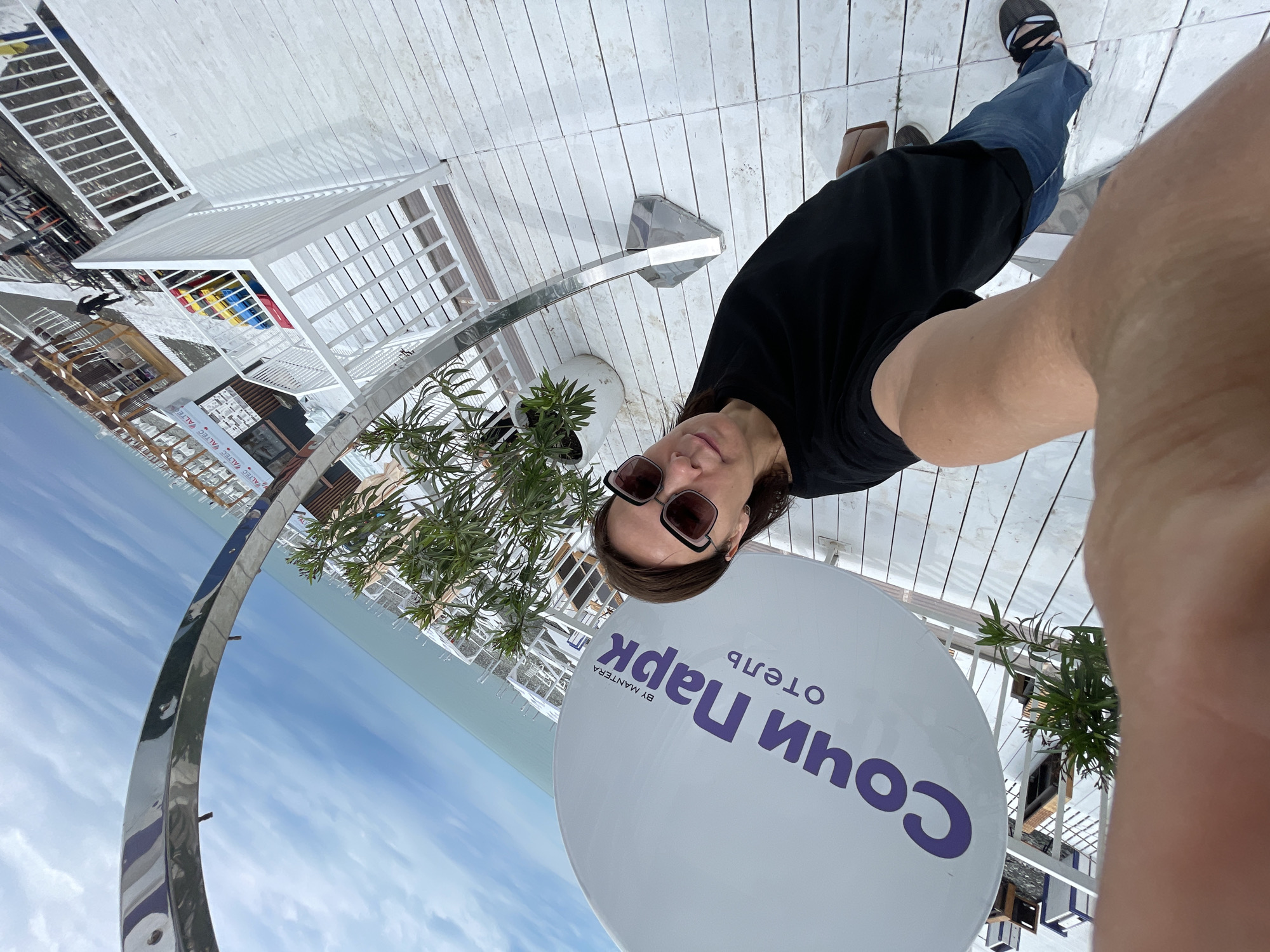 The image shows a selfie of a woman wearing sunglasses, a black t-shirt, and jeans. She is standing on a white tiled surface. In the background is a sign that reads "Сочи Парк отель BY MANTERA," indicating a location in Sochi, Russia. Other elements include a metal arch, plants in white pots, and partial views of white metal structures. The background includes a glimpse of the sky and sea.