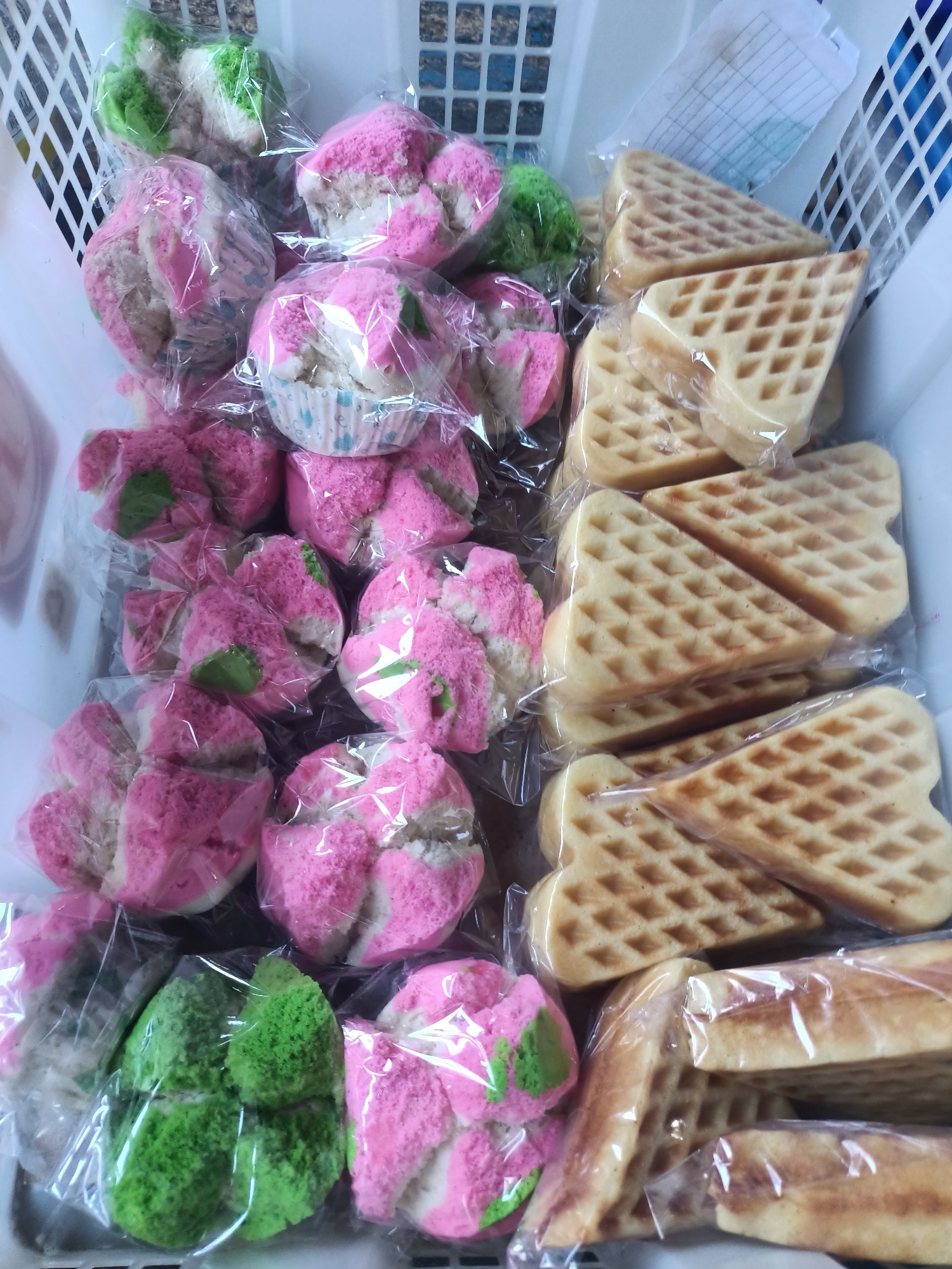 In Badas, Indonesia, a white plastic basket overflows with a variety of individually packaged baked goods. Prominently featured are several brightly colored steamed cakes, known as "kue putu ayu," with swirls of pink and green on a white base. These cakes have a distinctive flower-like shape and are encased in transparent plastic. Alongside them are triangular waffles, golden brown and showing the characteristic grid pattern of their baking molds, also individually wrapped. Some of the waffles appear to be stacked, while others are layered individually within the basket. The background shows the textured pattern of the plastic basket and a glimpse of a white paper document tucked into the upper right corner. The lighting suggests an indoor setting, likely a marketplace or a home kitchen where these treats are prepared or sold.