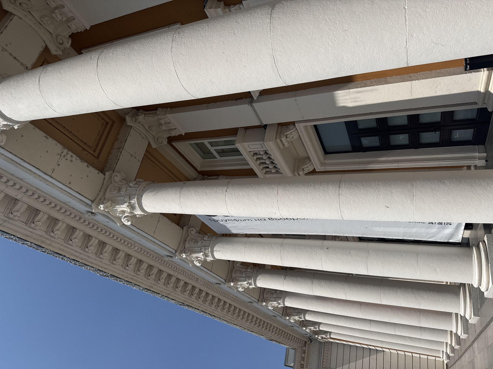 The image shows a low-angle view of the facade of the Palace of Culture in Pernik, Bulgaria, under a clear blue sky. A series of large, fluted white columns with Corinthian capitals line the front of the building, creating a colonnade. Between the columns, a large white banner with black text, appearing to be in Cyrillic script, hangs down. The building's exterior wall is painted a warm orange-brown color. To the right, a dark, multi-paned double door is visible, with a small white balcony above it. The architecture is neoclassical. There are no people visible in the frame. The ground in front of the building is paved with light-colored tiles.