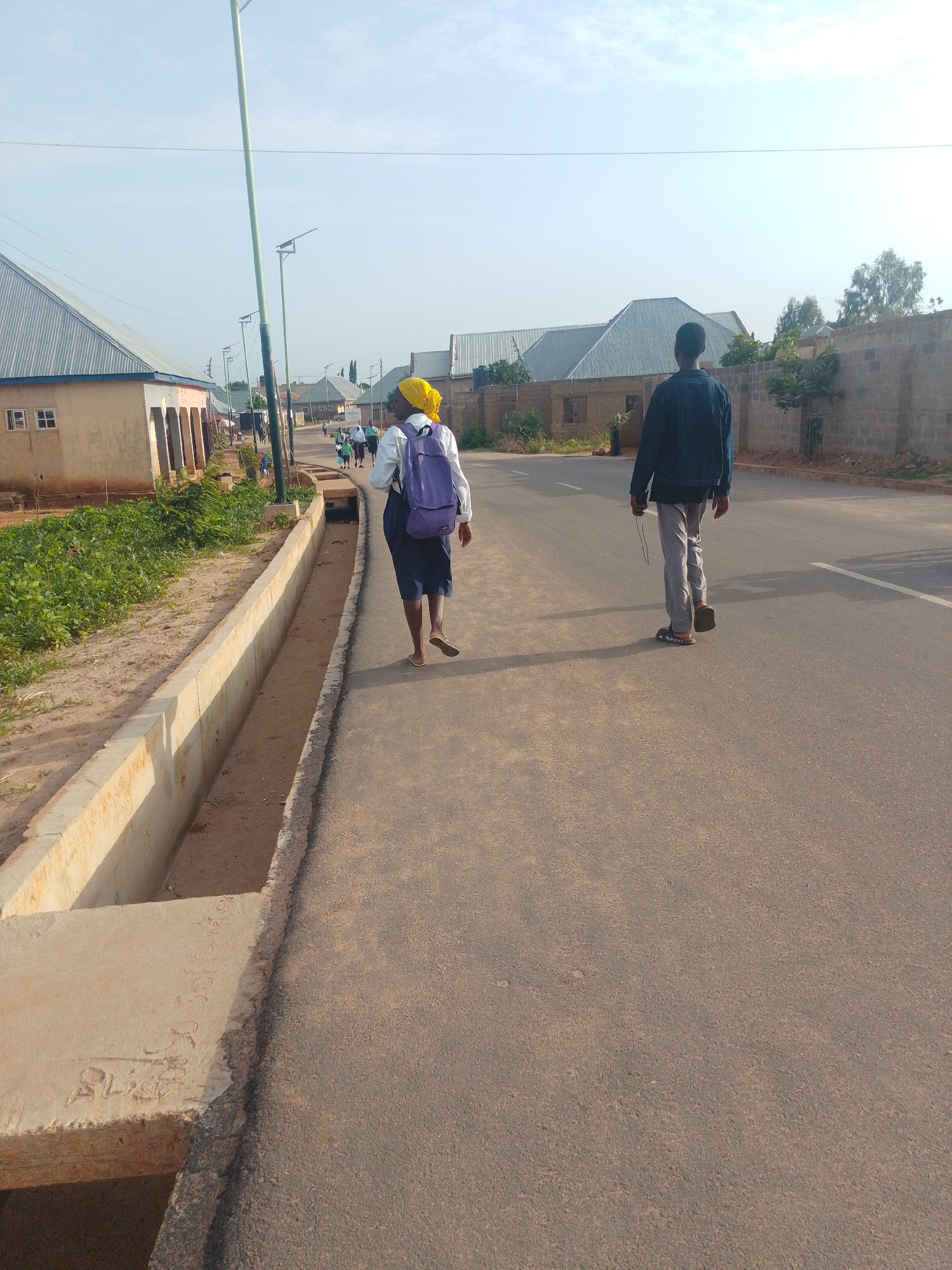 In Tempure, Nigeria, two individuals walk away from the camera on a paved road. On the left side of the road, a drainage ditch runs alongside a sandy area with some vegetation. Beyond the ditch, there are several single-story buildings with corrugated metal roofs and beige walls. The buildings appear to be residential or small commercial establishments. A street light with a solar panel is visible on the left side of the road. In the distance, more buildings and trees can be seen. On the right side of the road, a brick wall runs along the property line, with trees behind it. The sky is clear and bright. The person on the left is wearing a yellow headscarf, a white shirt, a blue backpack, and a dark skirt. The person on the right is wearing a dark denim jacket, light gray pants, and dark sandals. The road has a white line marking the center.