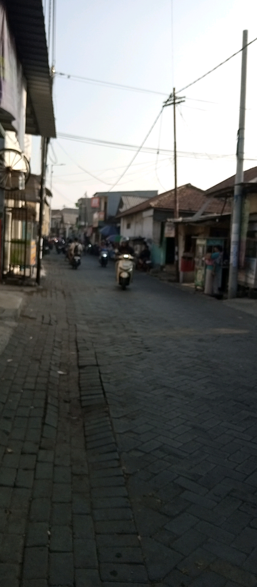 The image depicts a street scene in Wadungasri, Indonesia, likely during the late afternoon or early evening based on the lighting. The street is paved with dark, rectangular bricks laid in a herringbone pattern. On the left side of the street, there are buildings with corrugated metal roofs and awnings, and a metal fence. A decorative metal wheel is attached to the side of one of the buildings.
The street is populated with several motorbikes, some of which are moving. One motorbike is in the foreground, facing towards the viewer, with a rider wearing a helmet. Other motorbikes are visible further down the street, with riders.
On the right side of the street, there are more buildings, including what appear to be small shops or stalls with awnings. There are power lines and utility poles overhead. The sky is a pale, clear color, indicating a lack of significant cloud cover. The overall impression is of a busy, active street in a commercial or residential area.