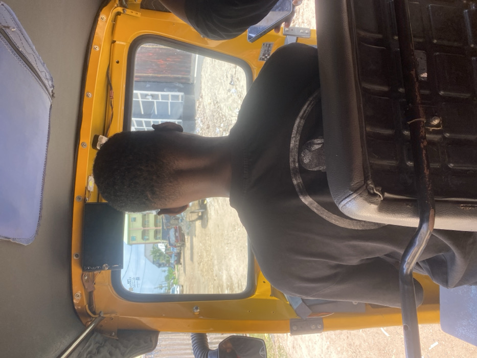 The image presents a forward-facing view from inside a yellow three-wheeled vehicle, likely a tricycle (keke napep), situated in Iyiowa Odekpa, Nigeria. The foreground is dominated by the back of an individual's head and upper torso, centrally positioned. This person has short, dark, closely-cropped hair and wears a dark-colored t-shirt, on the back of which a circular graphic or logo is partially visible. They are seated directly behind a black padded seat, which features a horizontal metal bar across its lower rear.
An electronic device, dark in color, is mounted above the vehicle's front windshield. To the right of the frame, a hand is partially visible, holding a smartphone with an illuminated screen.
Through the windshield, an unpaved, dusty road is visible, extending into the mid-ground. Several vehicles, including a light-colored sedan, are present on this road. On the left side of the road, a multi-story building with light-colored walls and green accents (possibly roofing or window frames) is discernible. To the immediate right, a darker-colored building with a ribbed or textured facade stands. The sky above is bright with some cloud cover, indicating daylight conditions, and sparse vegetation is visible on the horizon.