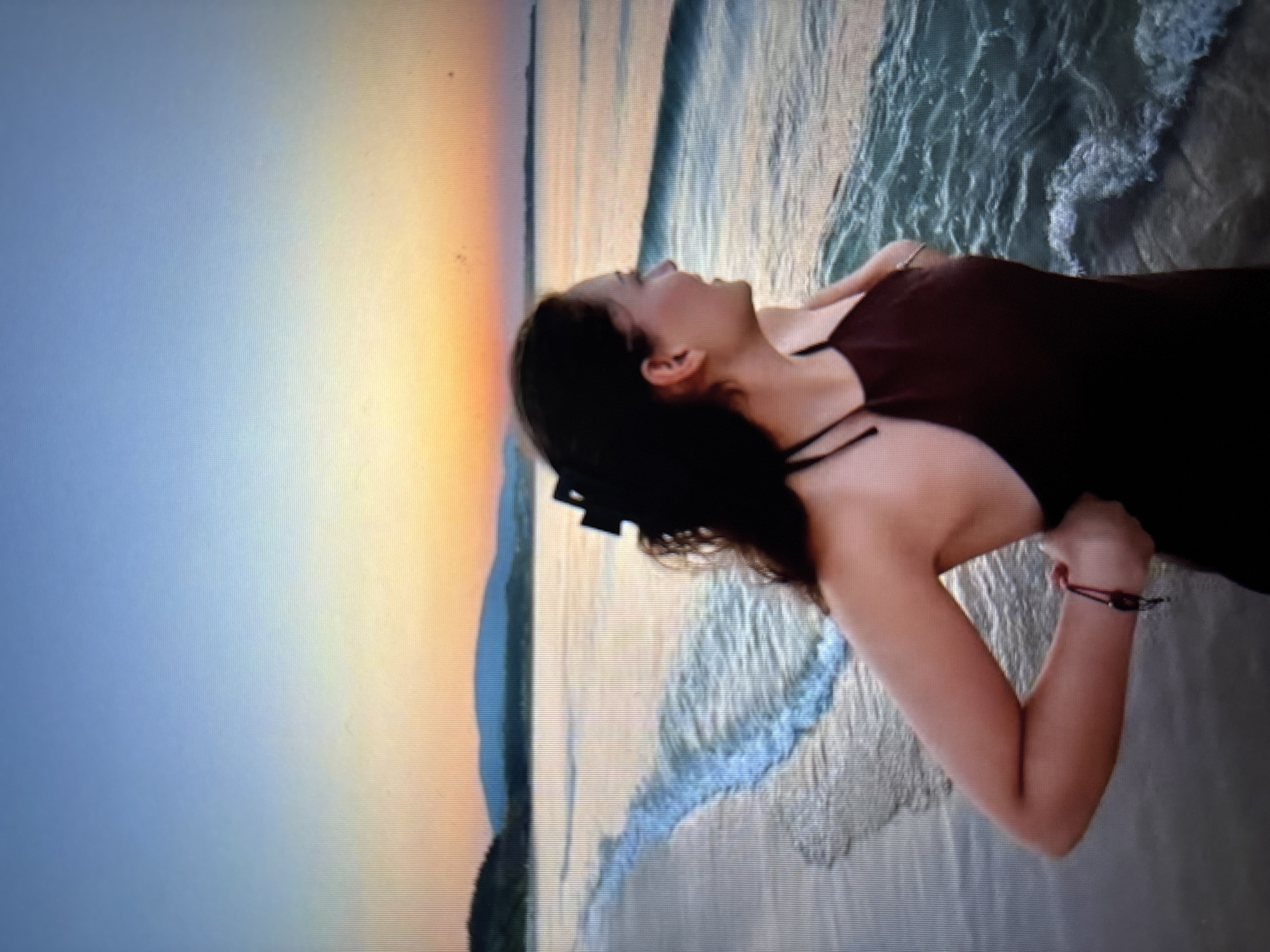 A young woman with dark hair pulled back stands on a beach looking out at the ocean as the sun sets. The sky transitions from a pale blue at the top to shades of orange and yellow near the horizon. The ocean waves are gentle, creating a thin line of foam at the shoreline. In the distance, there are rolling hills or mountains. The woman is wearing a dark, halter-neck dress and has a red string bracelet on her left wrist. The scene is peaceful and serene. This image was taken in Sofia, Bulgaria.